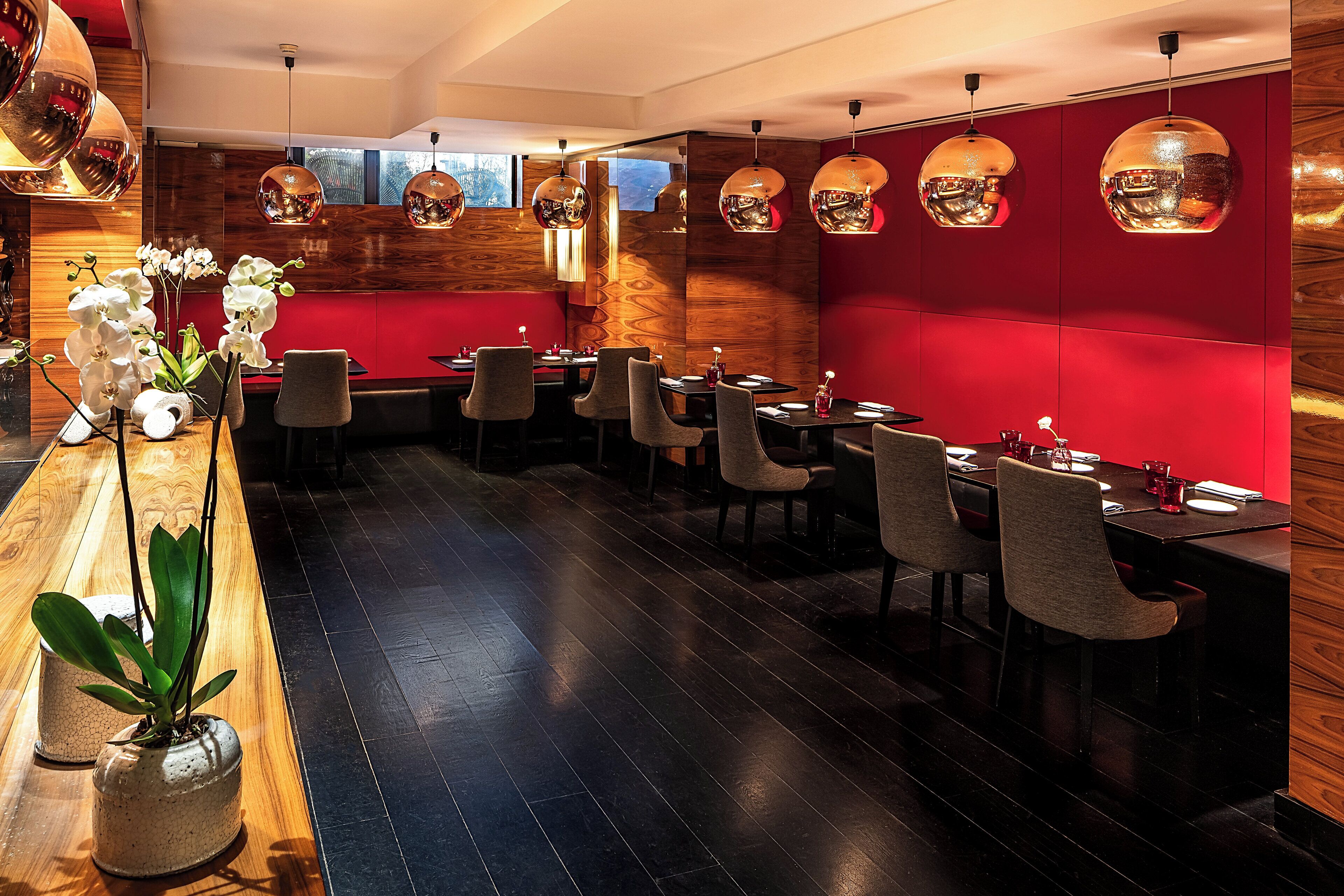 A modern restaurant interior with wooden flooring, red walls, and multiple dining tables with chairs.