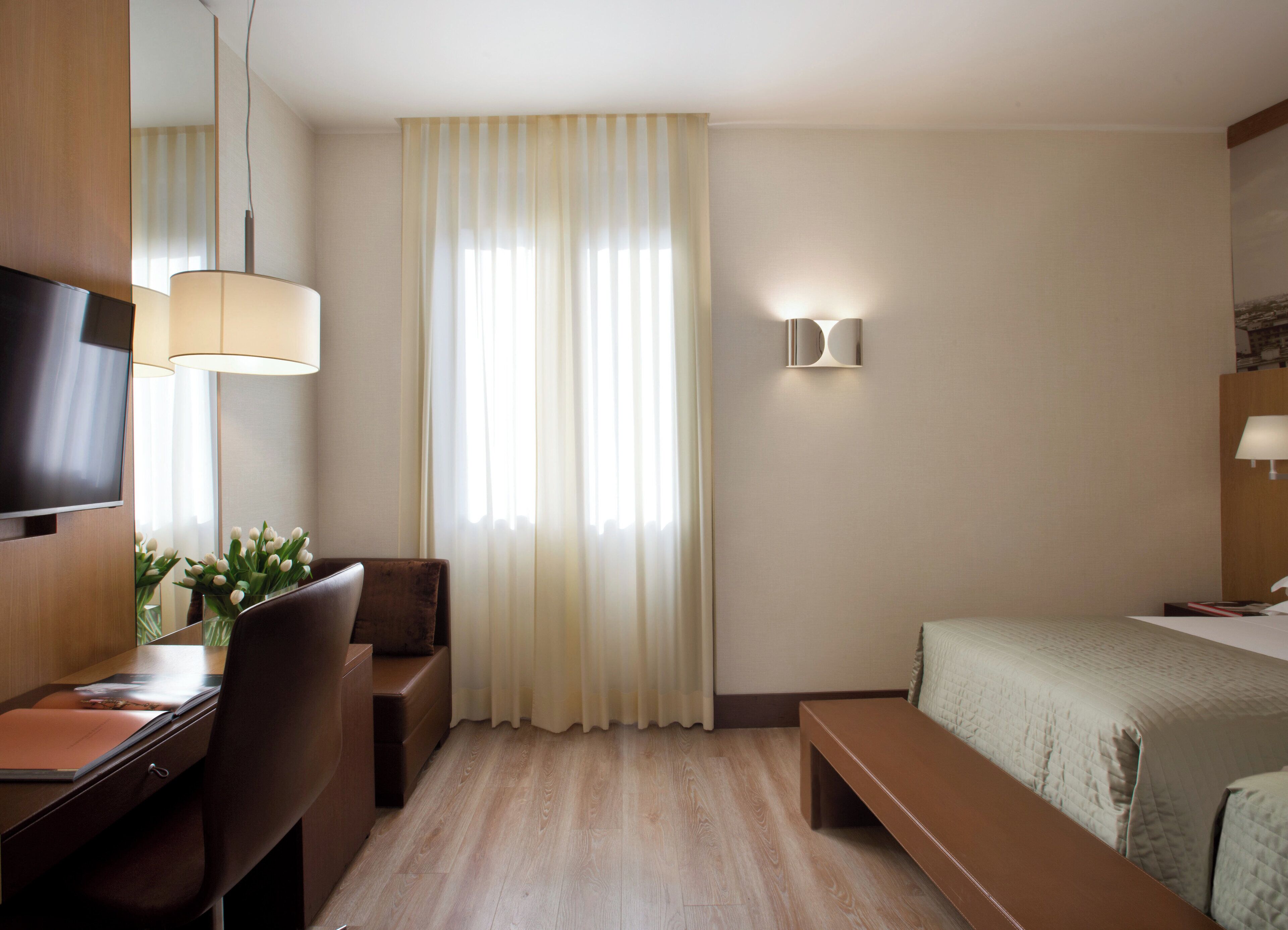 Hotellihuone, jossa on puinen työpöytä, tuoli, patalakanalla varustettu sänky, penkki, seinälle asennettu televisio ja ikkuna, jossa on verhot.