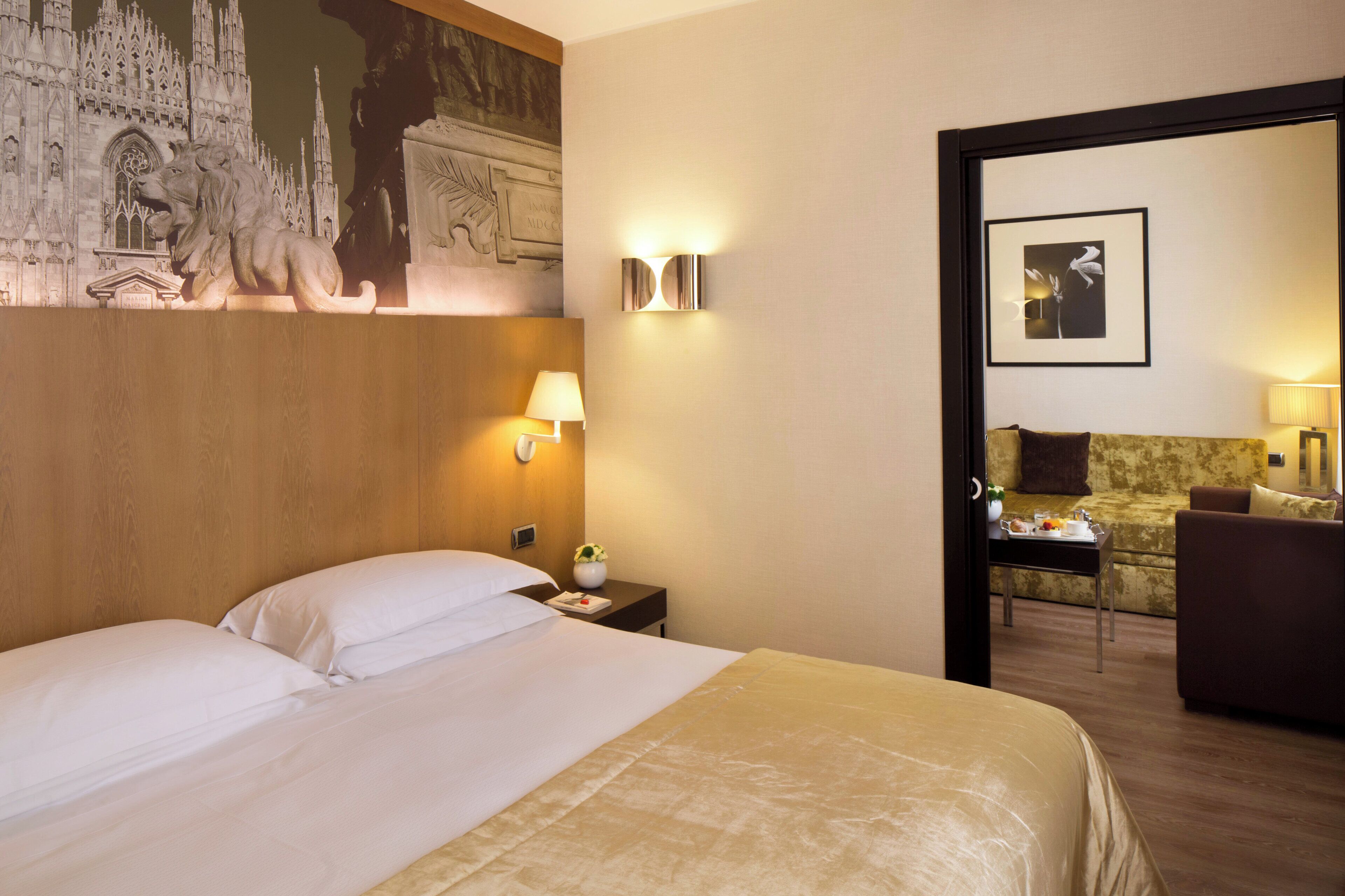 Habitación de hotel con una cama grande, una mesita de noche, una lámpara, una pequeña mesa con un florero y un sofá.