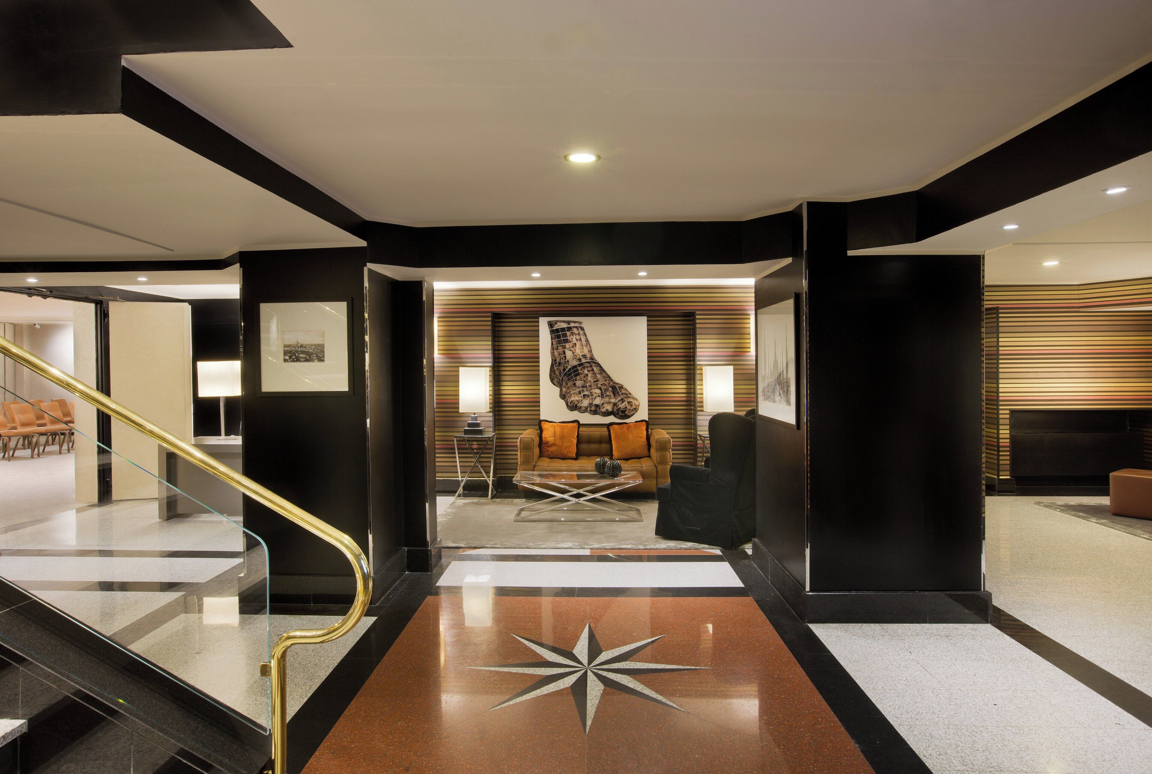 A modern lobby with a glass staircase, a central seating area with a sofa and coffee table, and a large abstract artwork on the wall.