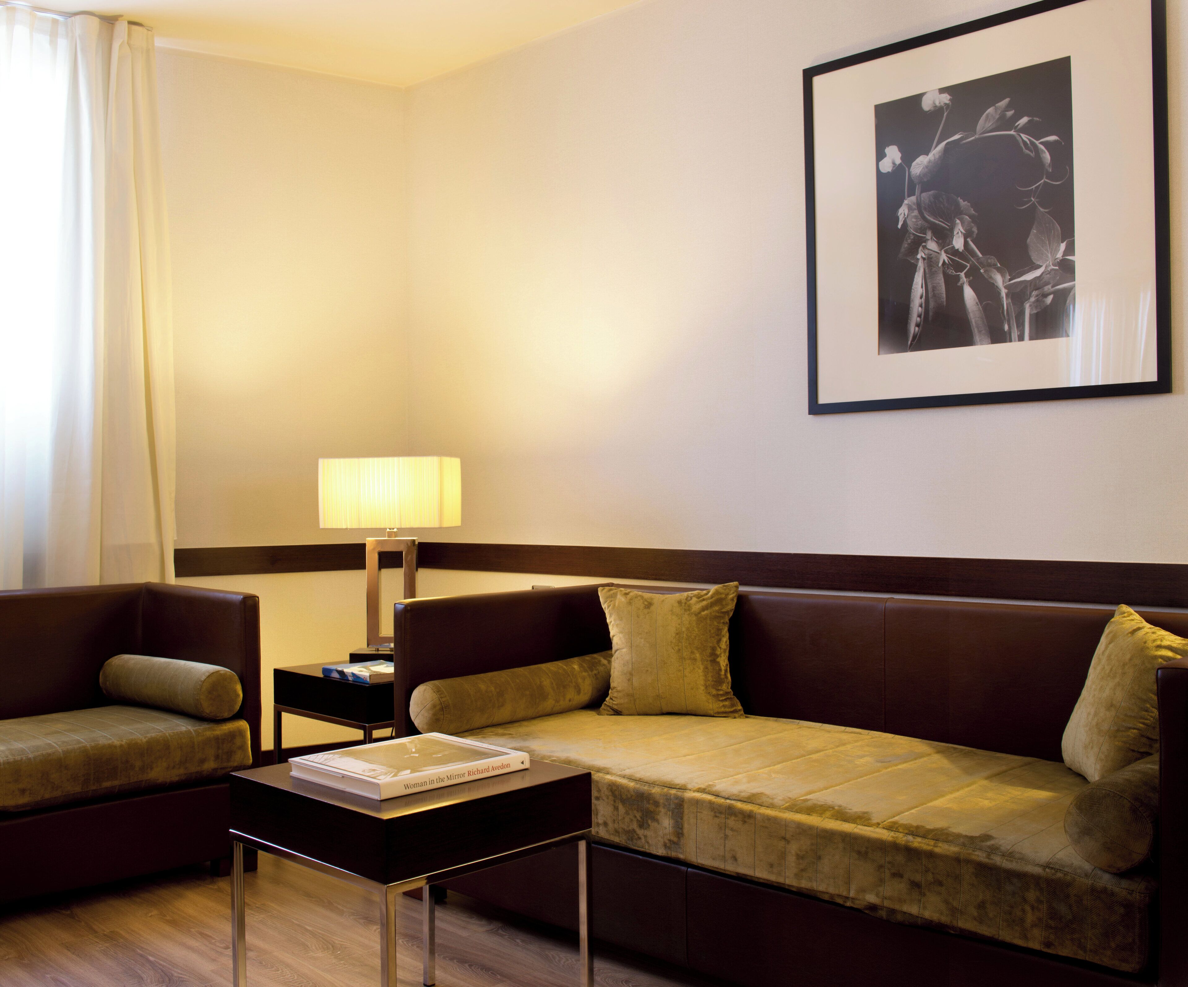 Una habitación de hotel con un sofá marrón, un sillón más pequeño, una mesita de noche con una lámpara y un cuadro en la pared.