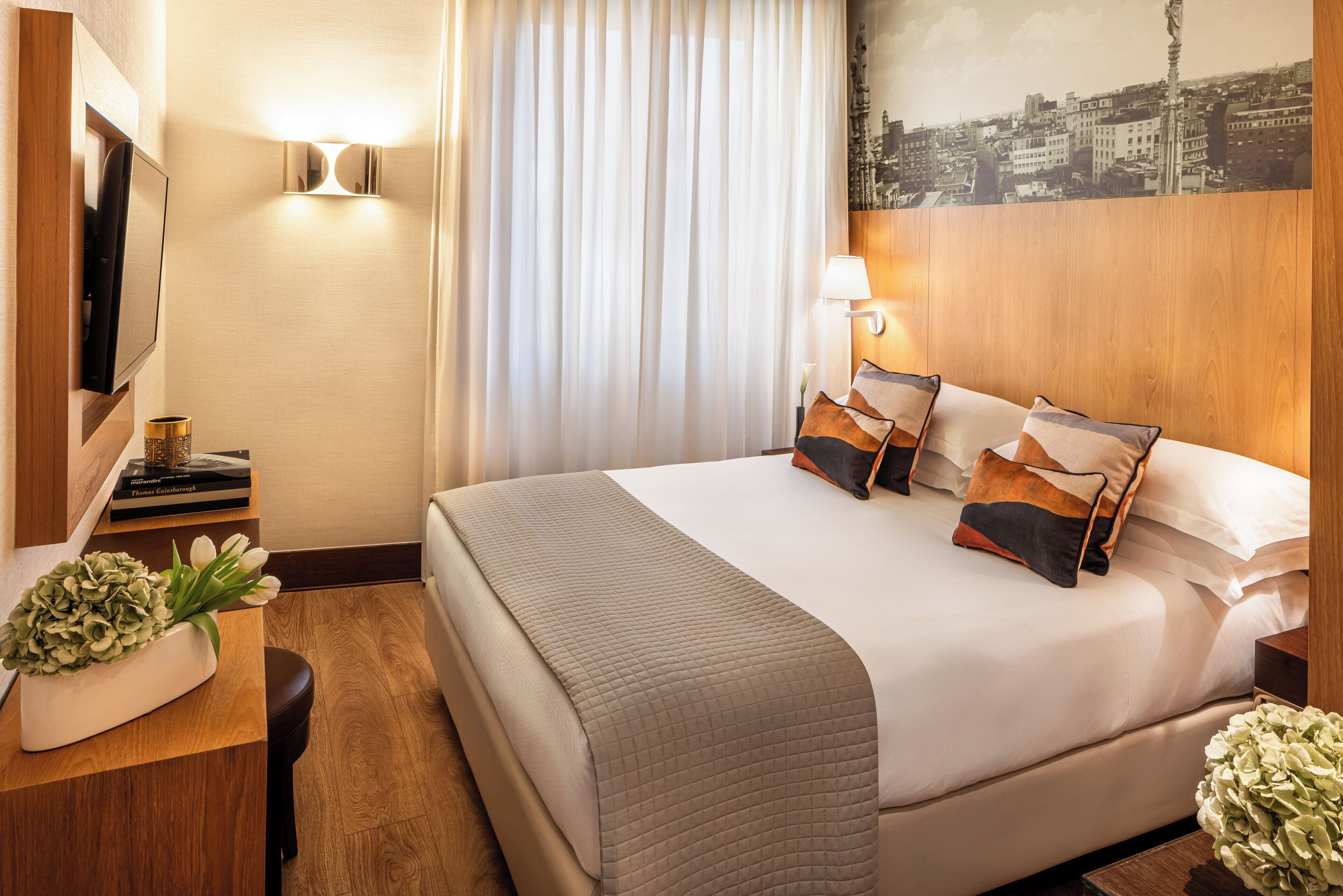 A hotel room with a large bed, a flat-screen TV, a desk with a lamp, and a cityscape painting on the wall.