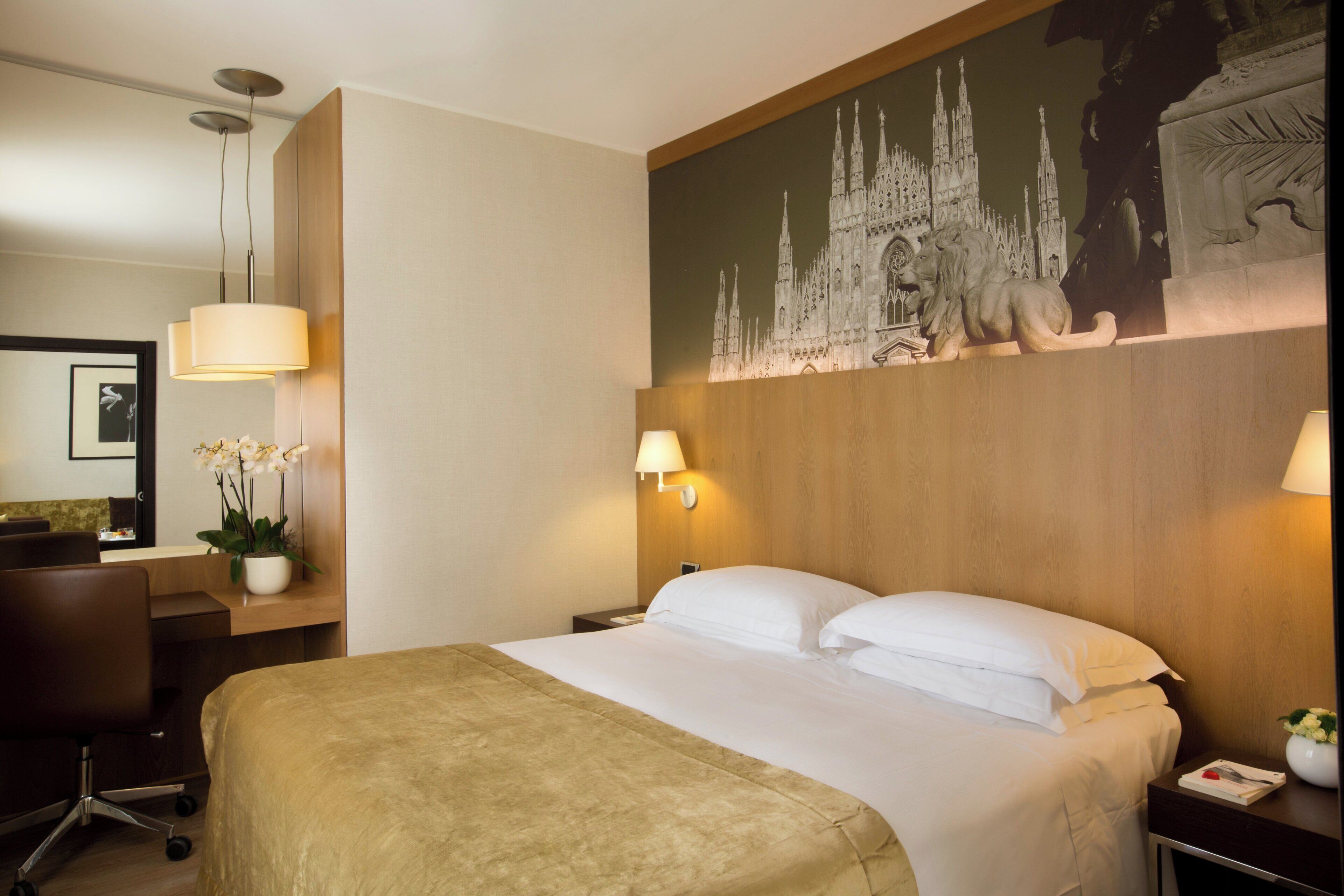 A hotel room with a large bed, a desk with a chair, a lamp, and a wall-mounted artwork of a castle.