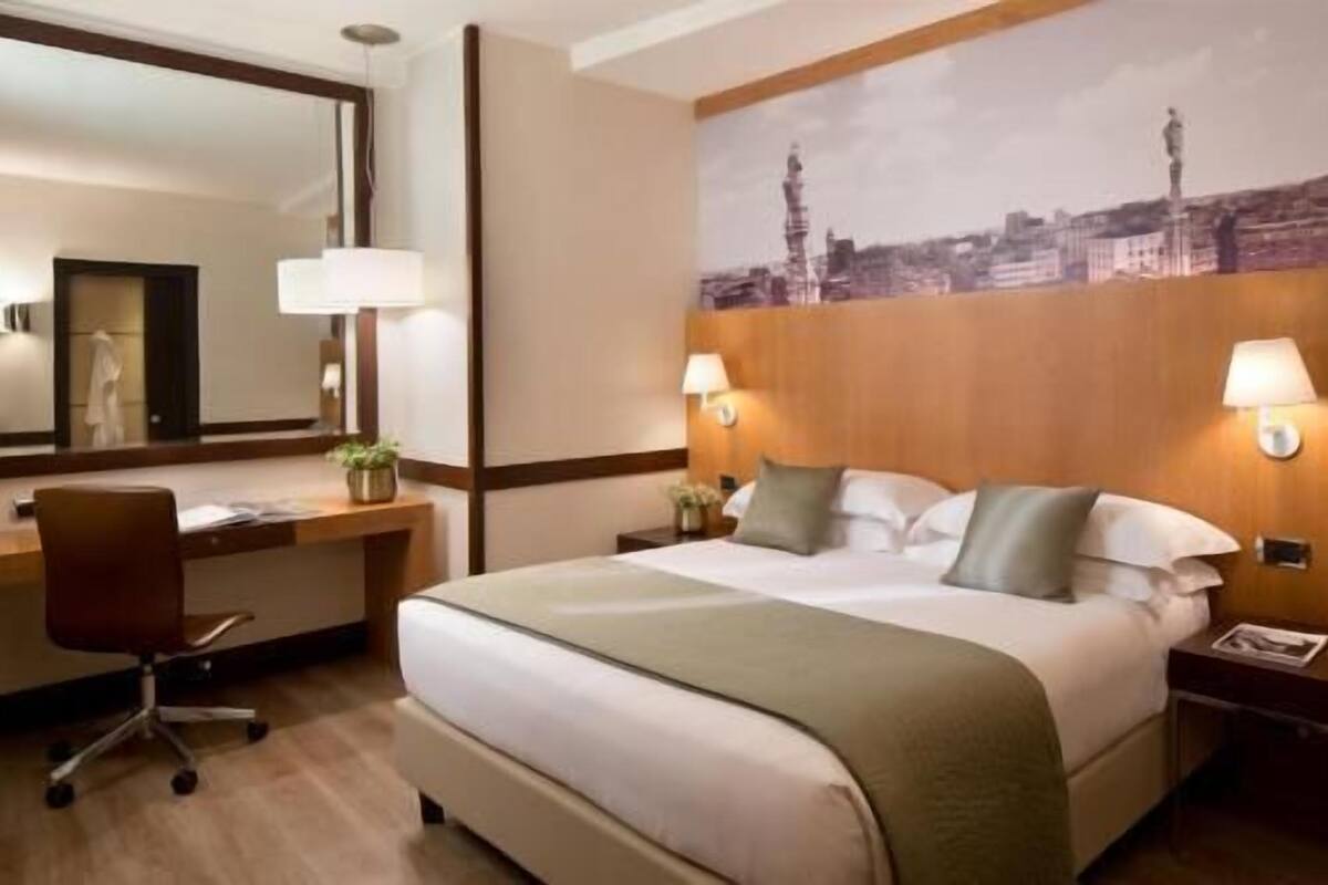 A hotel room with a large bed, a desk, a chair, and a wall-mounted lamp.