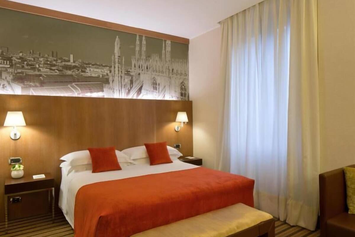 A hotel room with a large bed, two bedside lamps, a bench, a nightstand, and a cityscape painting on the wall.