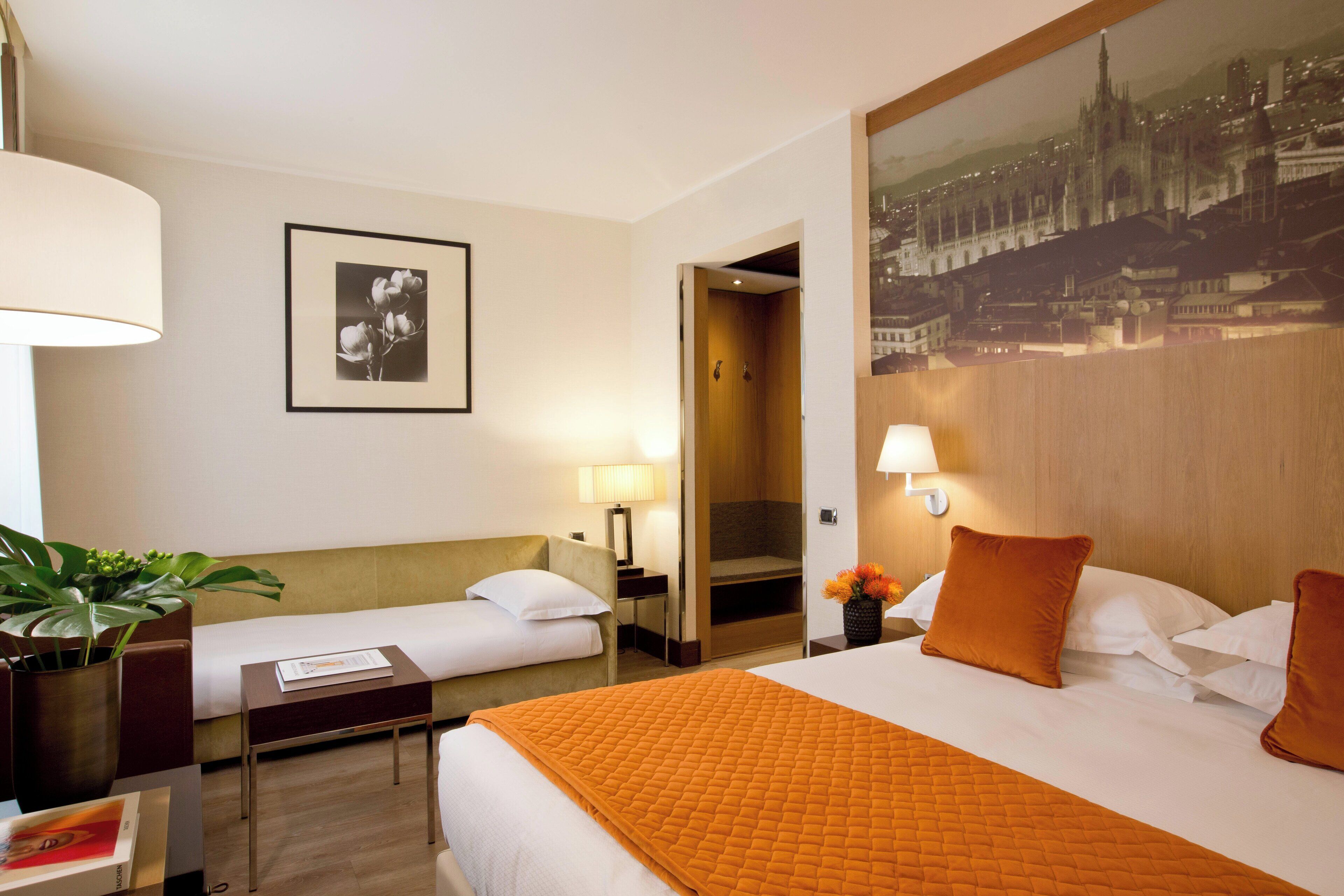 A hotel room with a large bed, a smaller bed, a desk, a chair, a lamp, a framed picture, and a potted plant.