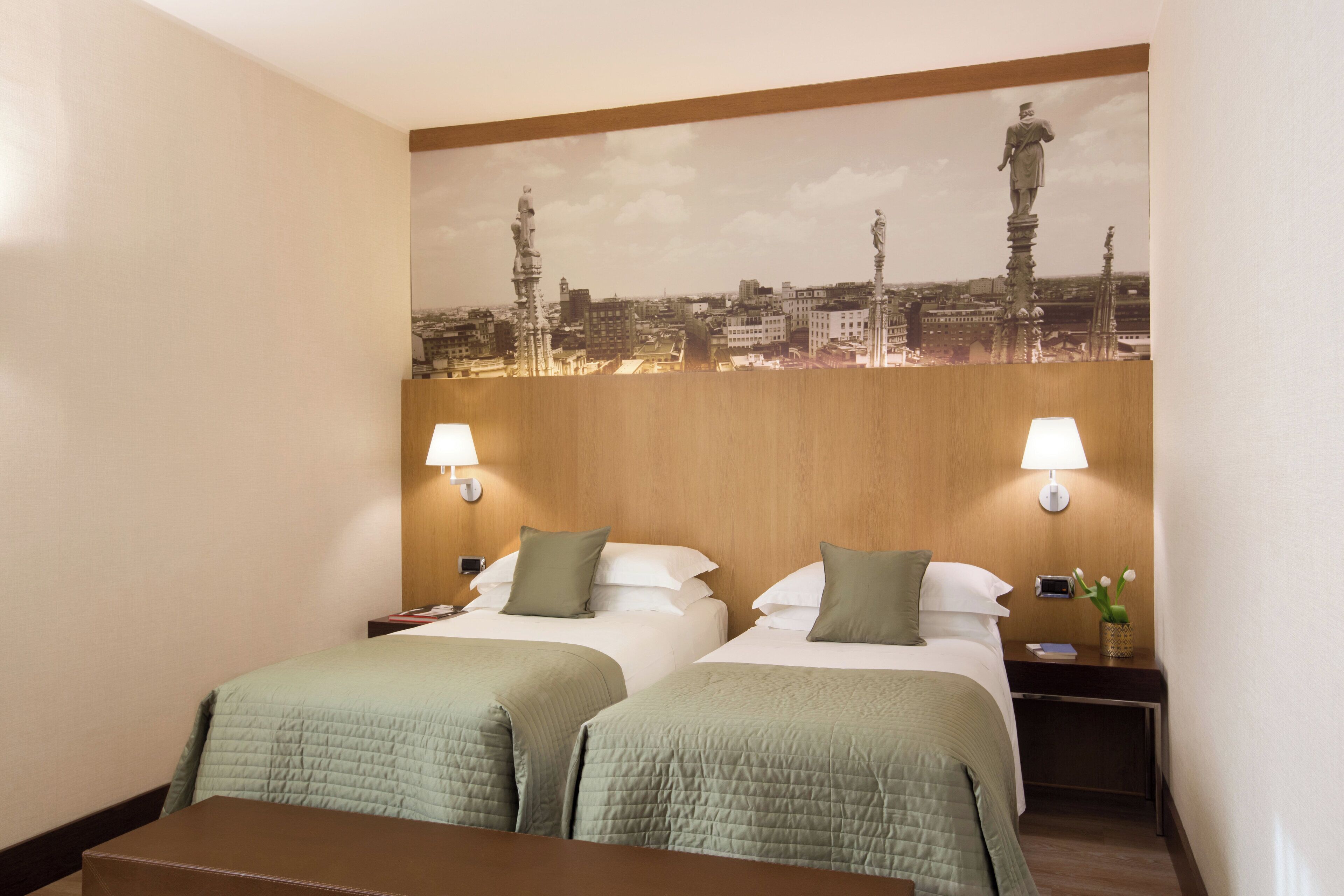 A hotel room with two beds, a wooden headboard, a cityscape mural, two bedside lamps, and a nightstand with a vase of flowers.