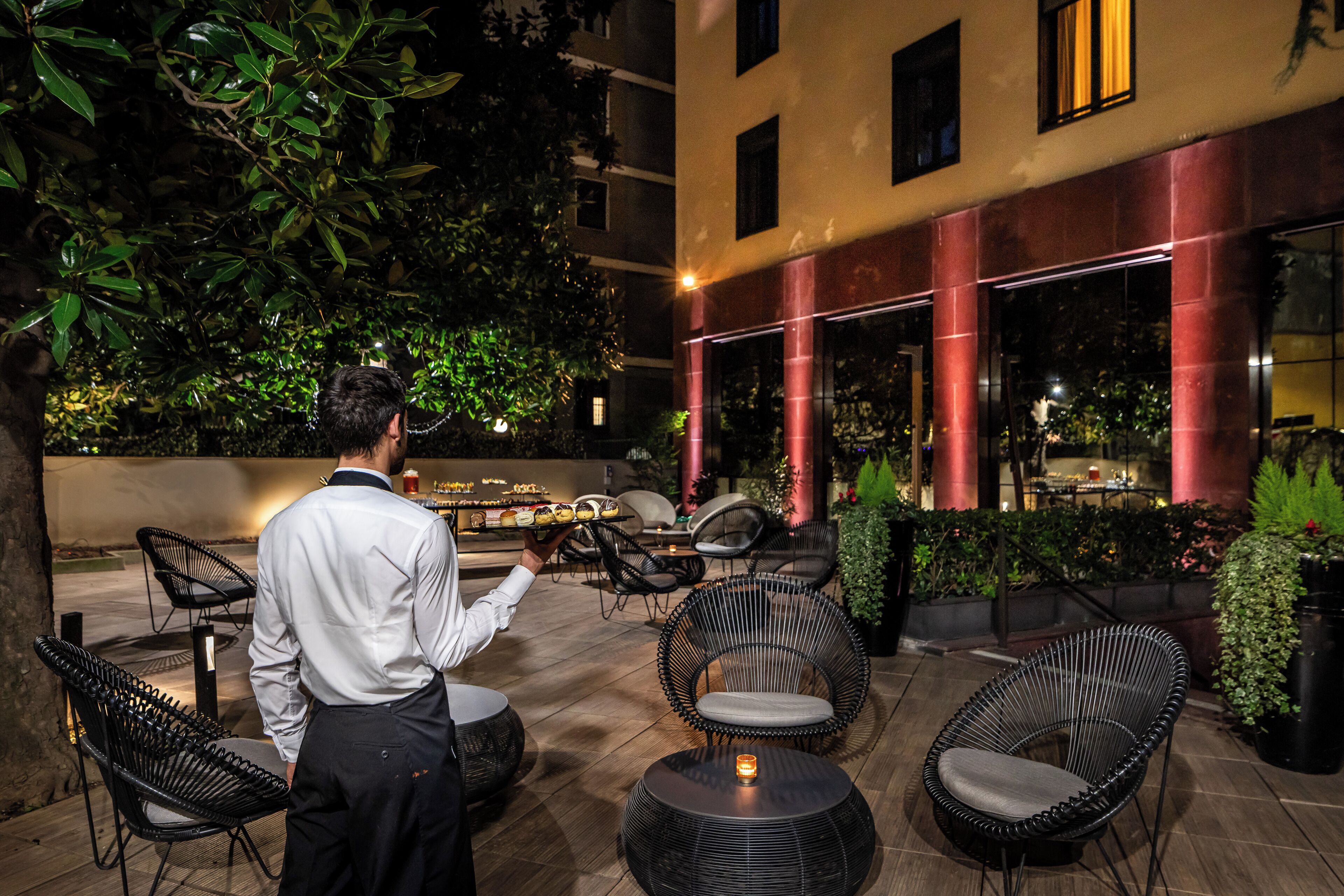 Un espace extérieur aménagé avec des chaises en osier et une petite table sur laquelle est posée une bougie, le tout entouré de verdure et adossé à un bâtiment aux piliers rouges.