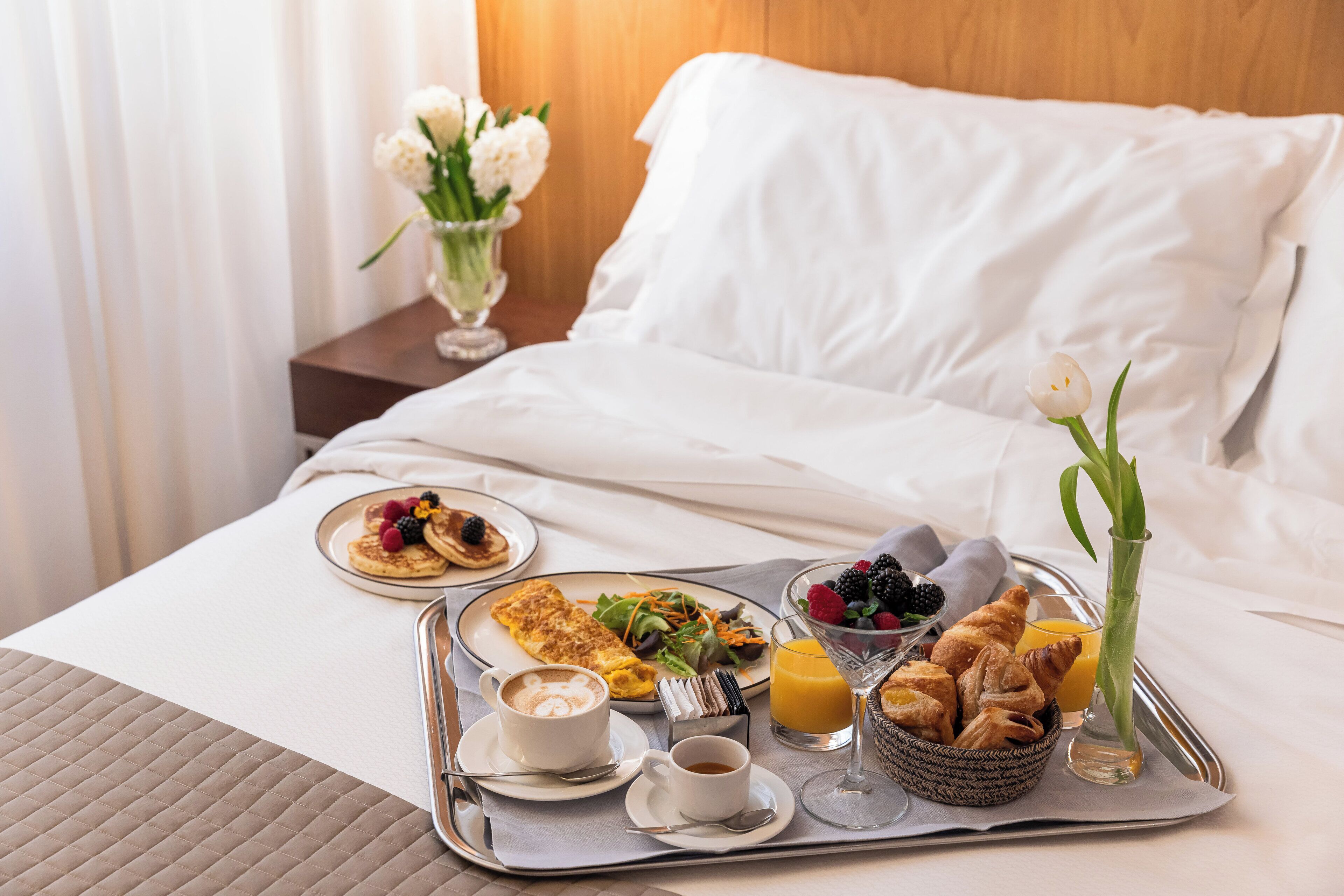 A neatly made breakfast tray on a bed with a white duvet, including pancakes, a croissant basket, coffee, and juice.