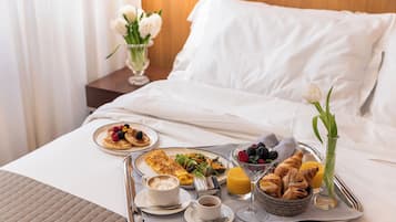 Un vassoio colazione ordinato su un letto con piumone bianco, con pancakes, cestino di croissant, caffè e succo di frutta.