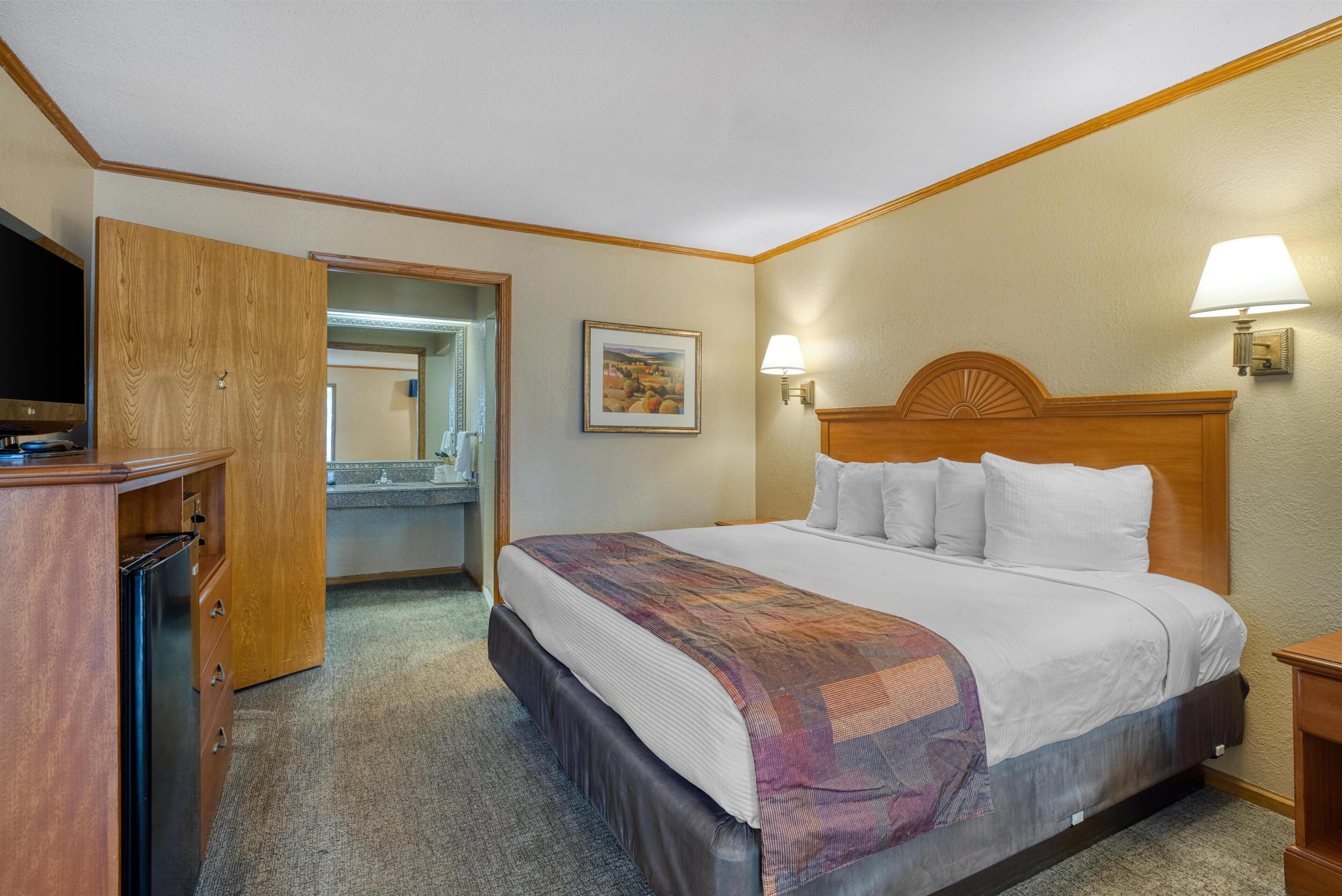 A hotel room with a bed, a wooden wardrobe, a mirror, a painting, and two wall-mounted lamps.