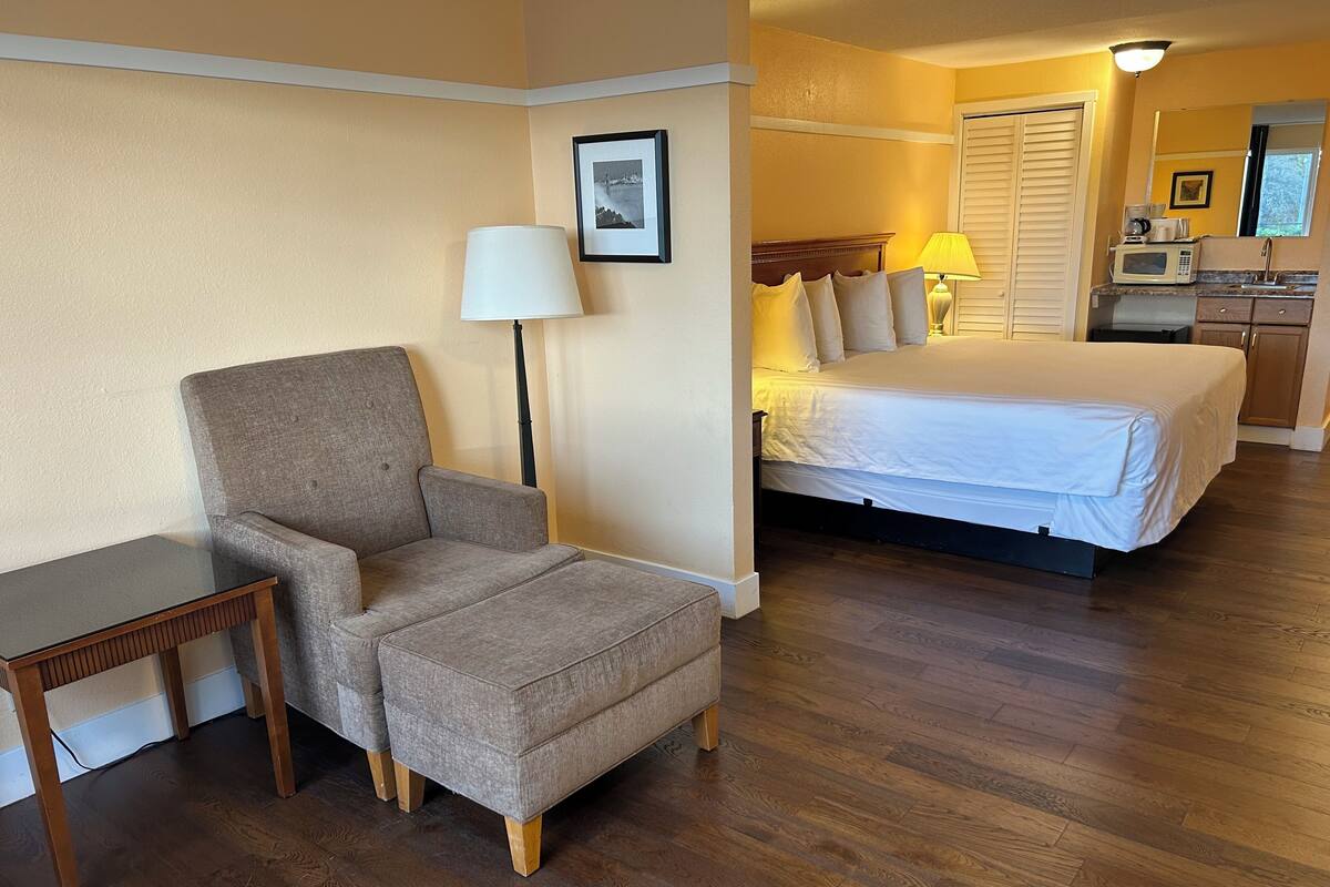 Quarto de hotel com cama, poltrona, mesinha, luminária e micro-ondas na kitchenette.