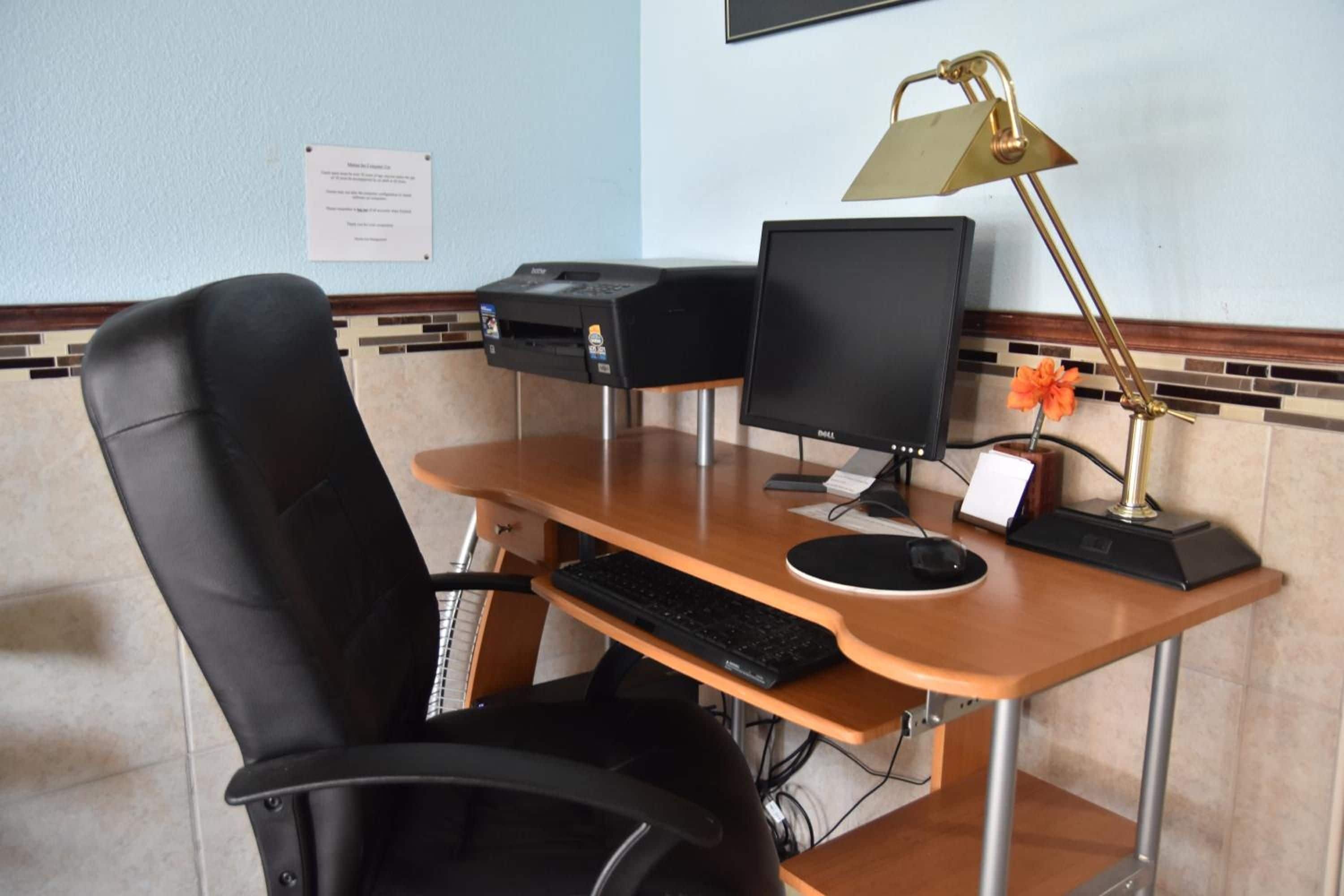 A hotel room with a wooden desk, a black office chair, a desktop computer, a printer, and a desk lamp.