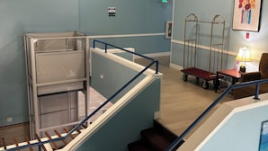 A hotel room with a blue wall, a wooden floor, a bed, a desk with a lamp, a chair, a cart, and a staircase. - Marina Inn (Des Moines)
