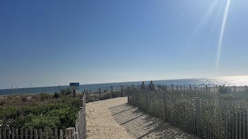 A sandy pathway leading to a beach with a wooden fence on one side and a signboard in the distance.