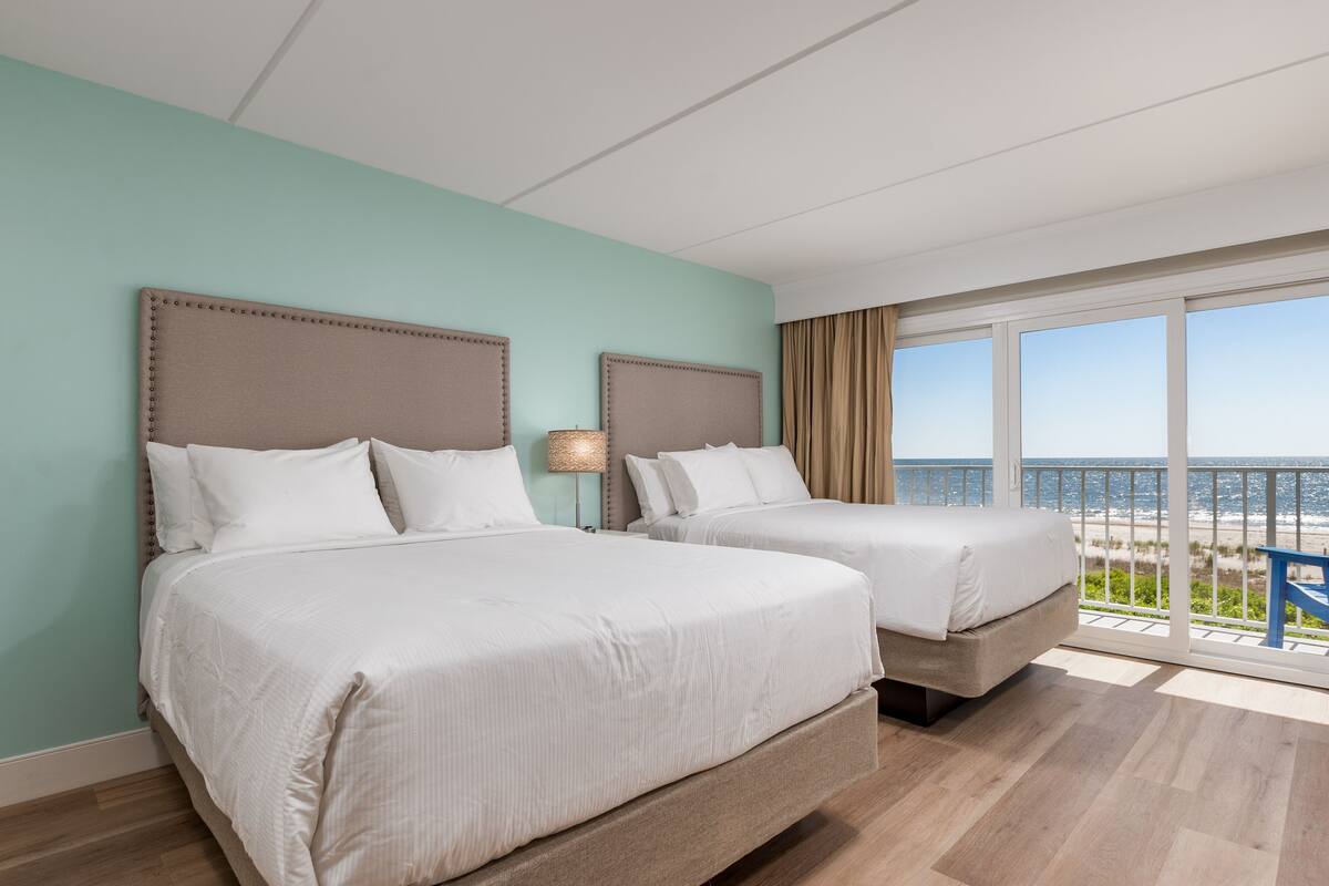 A hotel room with two beds, a large window with a view of the beach, and a balcony with a blue chair.
