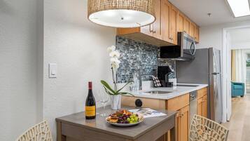 A modern kitchen with wooden cabinets, a microwave, and a refrigerator. There is a dining table with a bottle of wine, glasses, and a plate of fruit.
