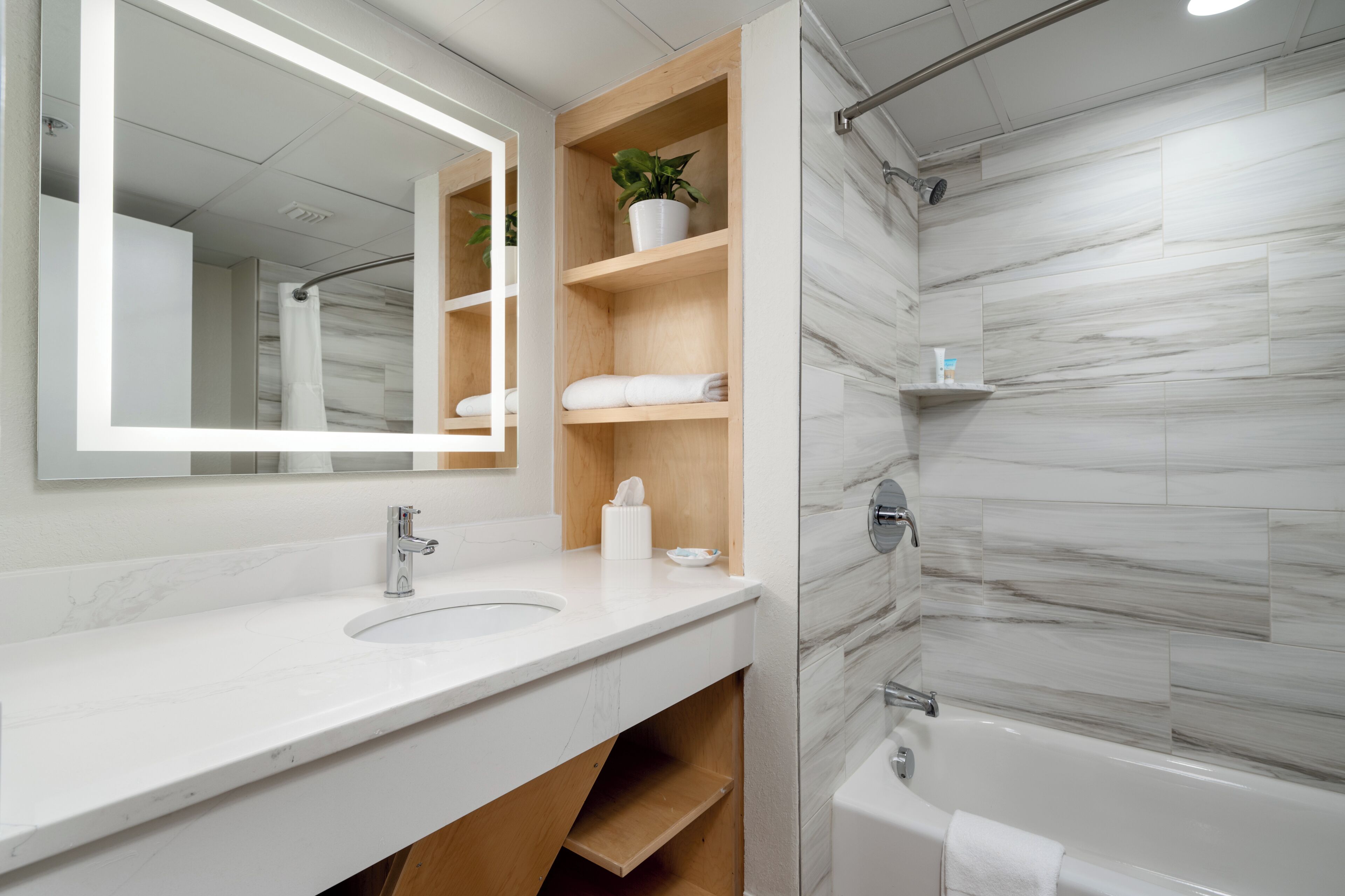 Un baño moderno con salpicadero de mármol, un espejo grande, un lavabo con grifo, zona de ducha y estantes de madera con toallas y una planta en maceta.