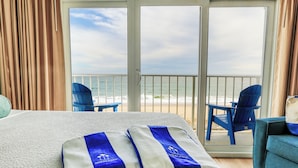 A hotel room with a bed, two blue chairs, a table, and a view of the beach.