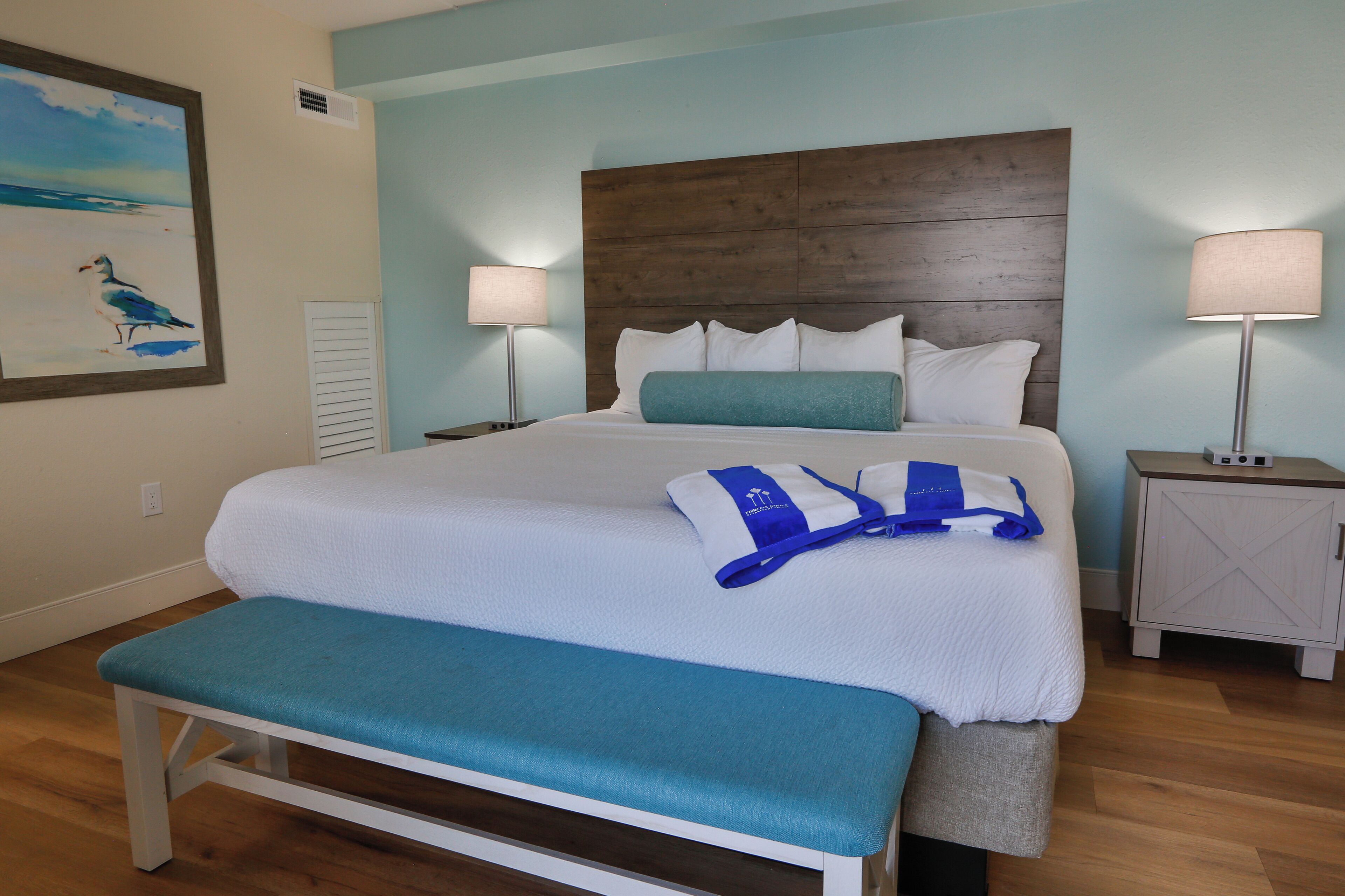 Una cama bien hecha con tablero de madera, dos lámparas de noche, un banco y un cuadro de un pájaro en la pared.