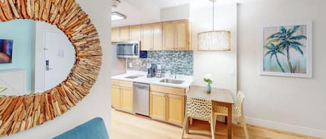 Een moderne keuken met houten kastjes, een magnetron en een spoelbak. Er is een eethoek met een tafel en stoelen, en een decoratieve spiegel aan de muur.