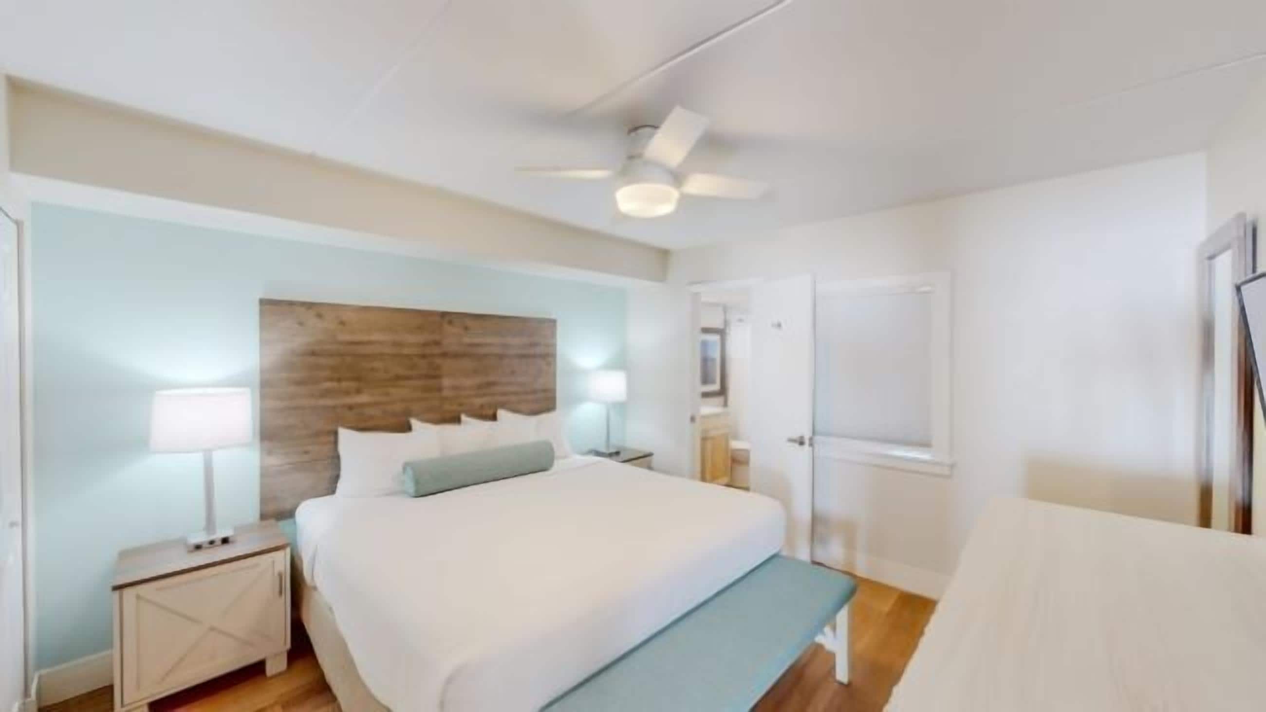 Een slaapkamer met een bed, nachtkastjes, een bankje, een plafondventilator en een deur.