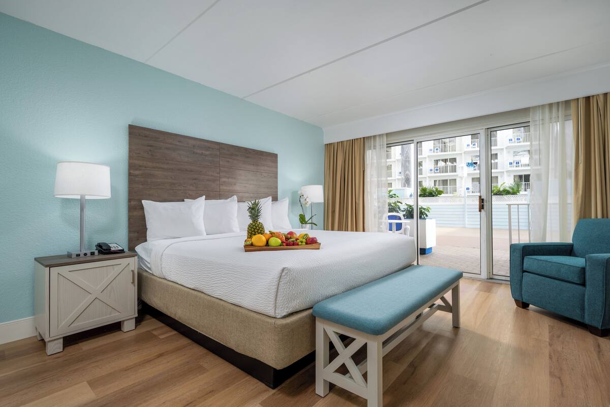 A hotel room with a bed, a bedside table with a lamp, a bench, a blue armchair, and a balcony with a view of buildings.