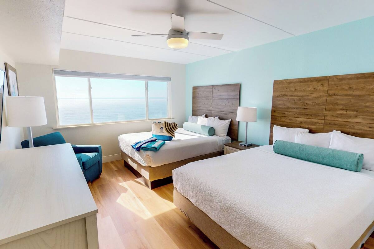 A hotel room with a large bed, a smaller bed, a desk, and a chair. There is a window with a view of the ocean.