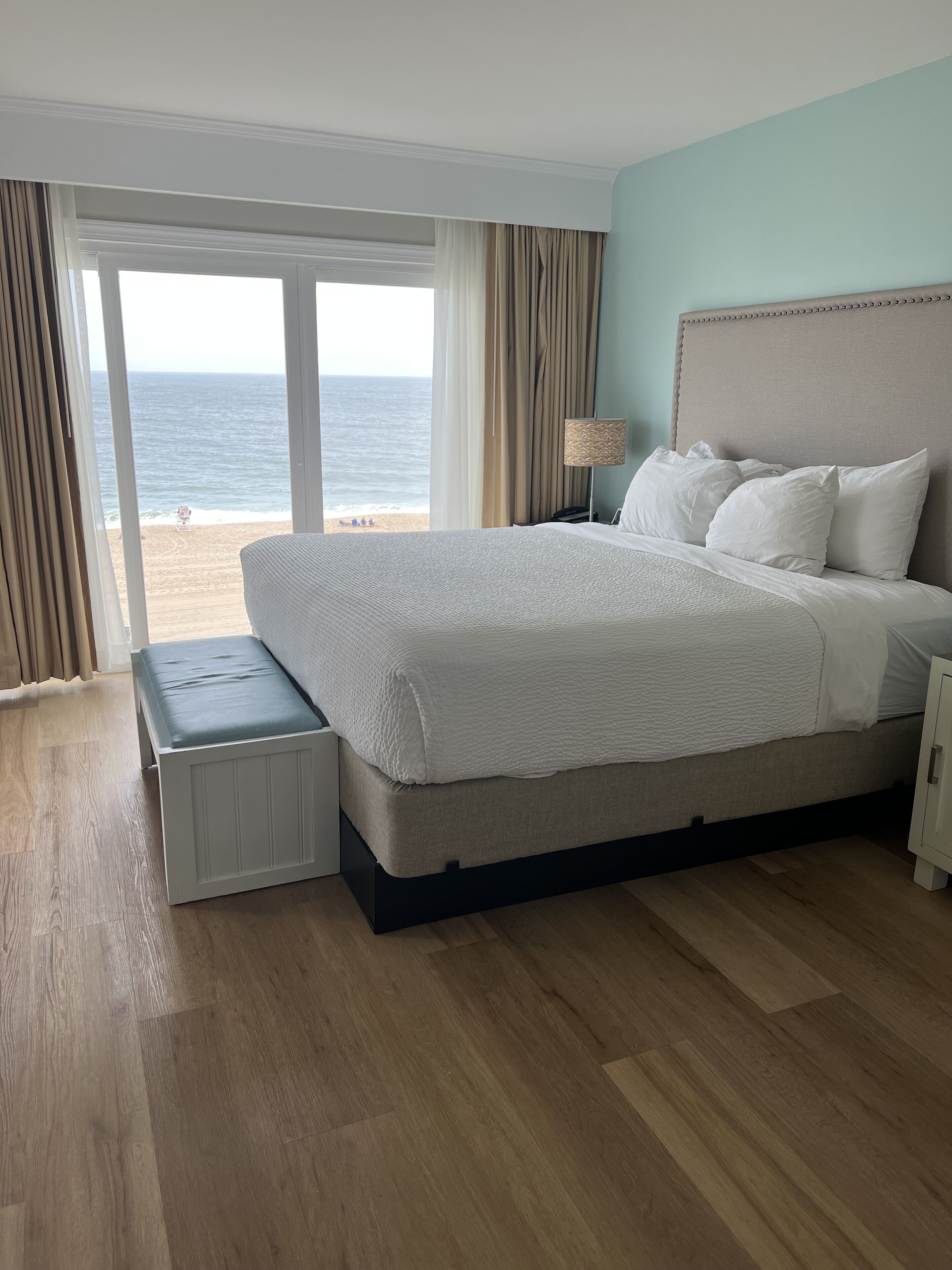 A bedroom with a large bed, a bench, a nightstand, and a view of the beach.