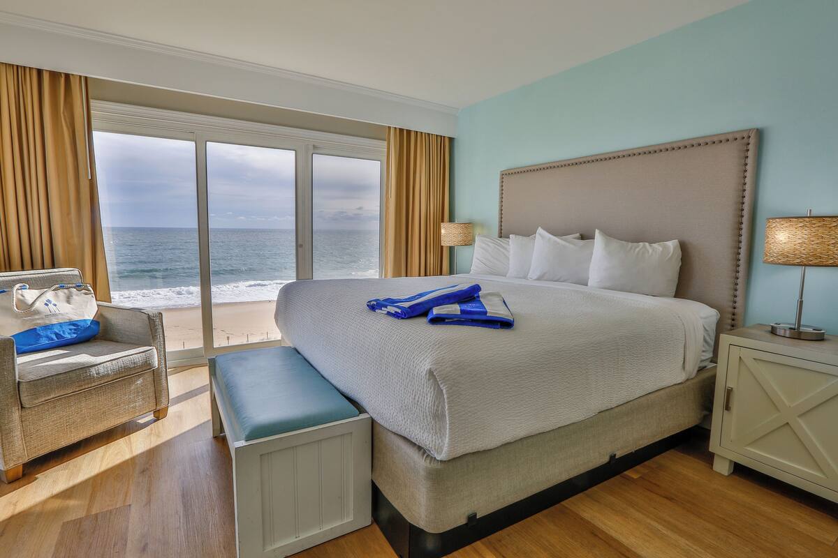 A hotel room with a large bed, a bench, a nightstand, and a view of the ocean.