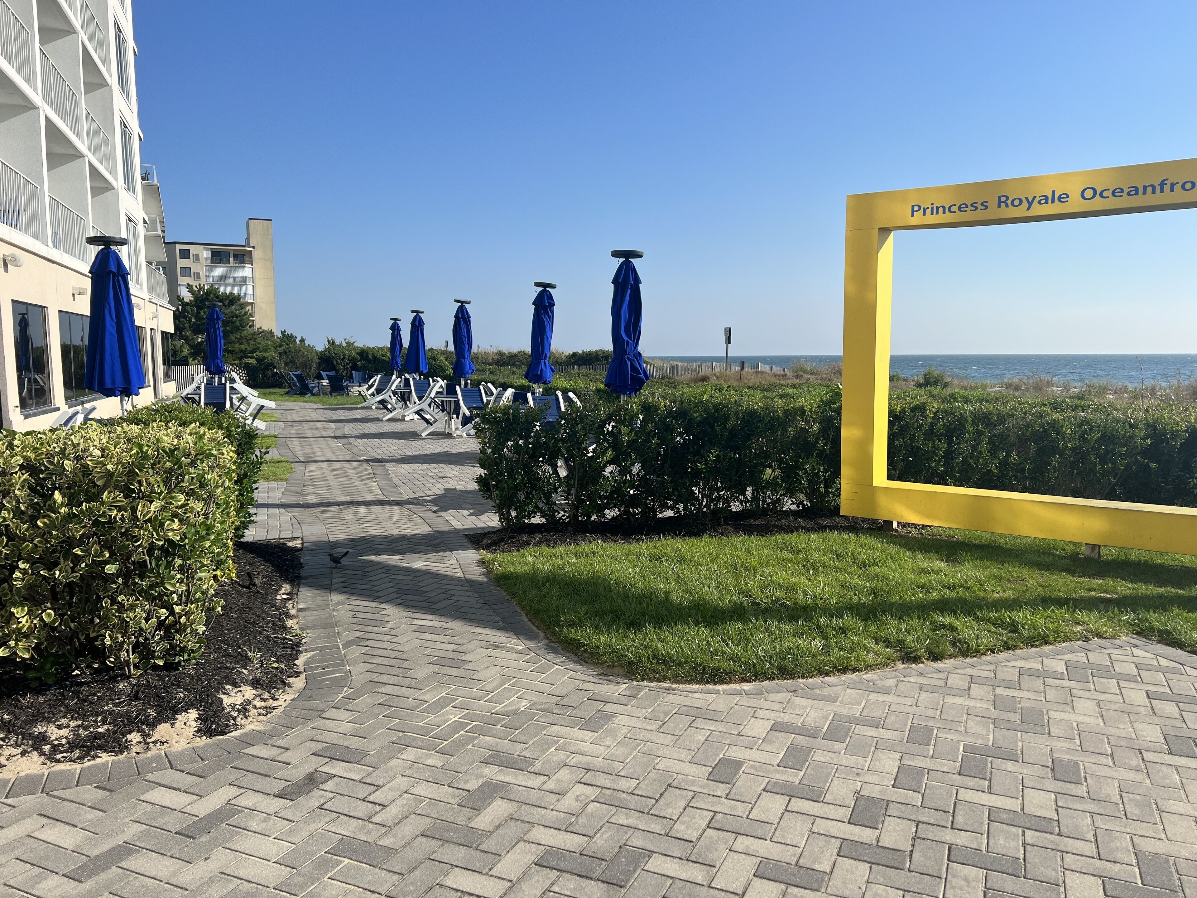 Un chemin pavé menant aux parasols de plage, un panneau jaune indiquant « Princess Royale Oceanfront », et une vue sur l’océan.