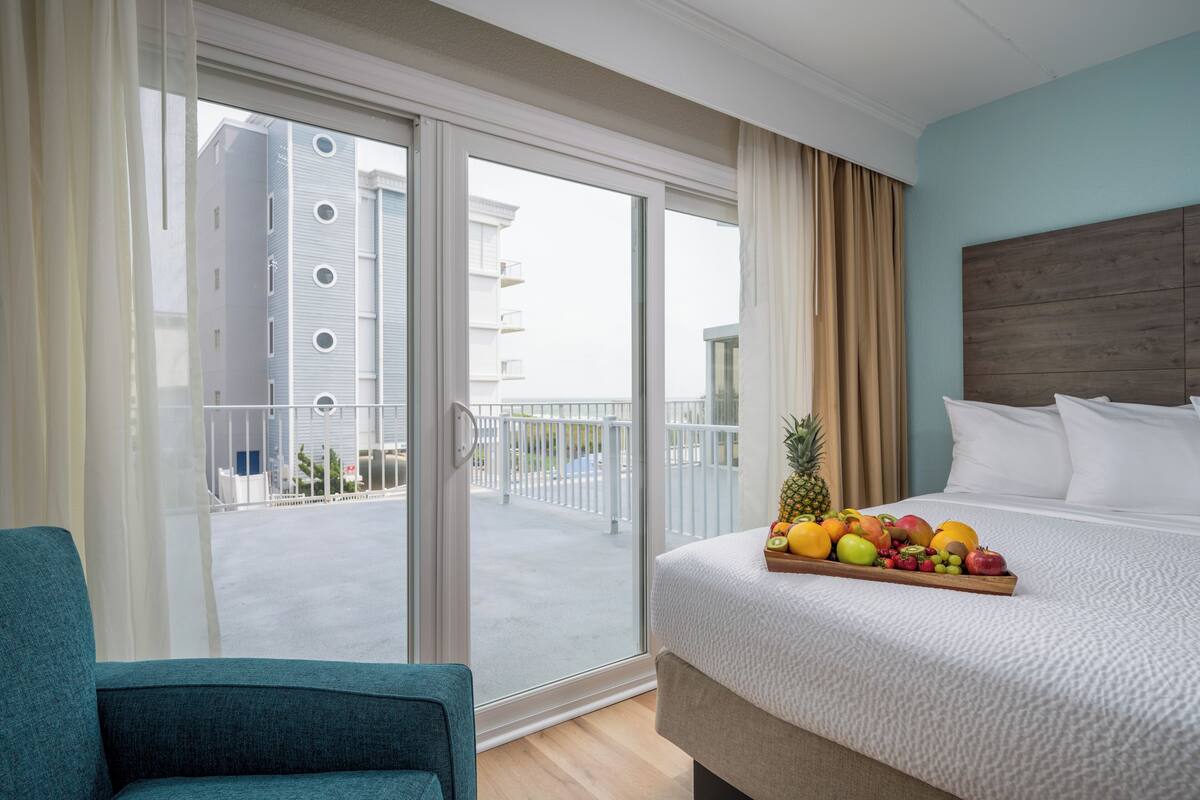 A hotel room with a bed, a fruit tray, a view of a building through a glass door, and a chair.