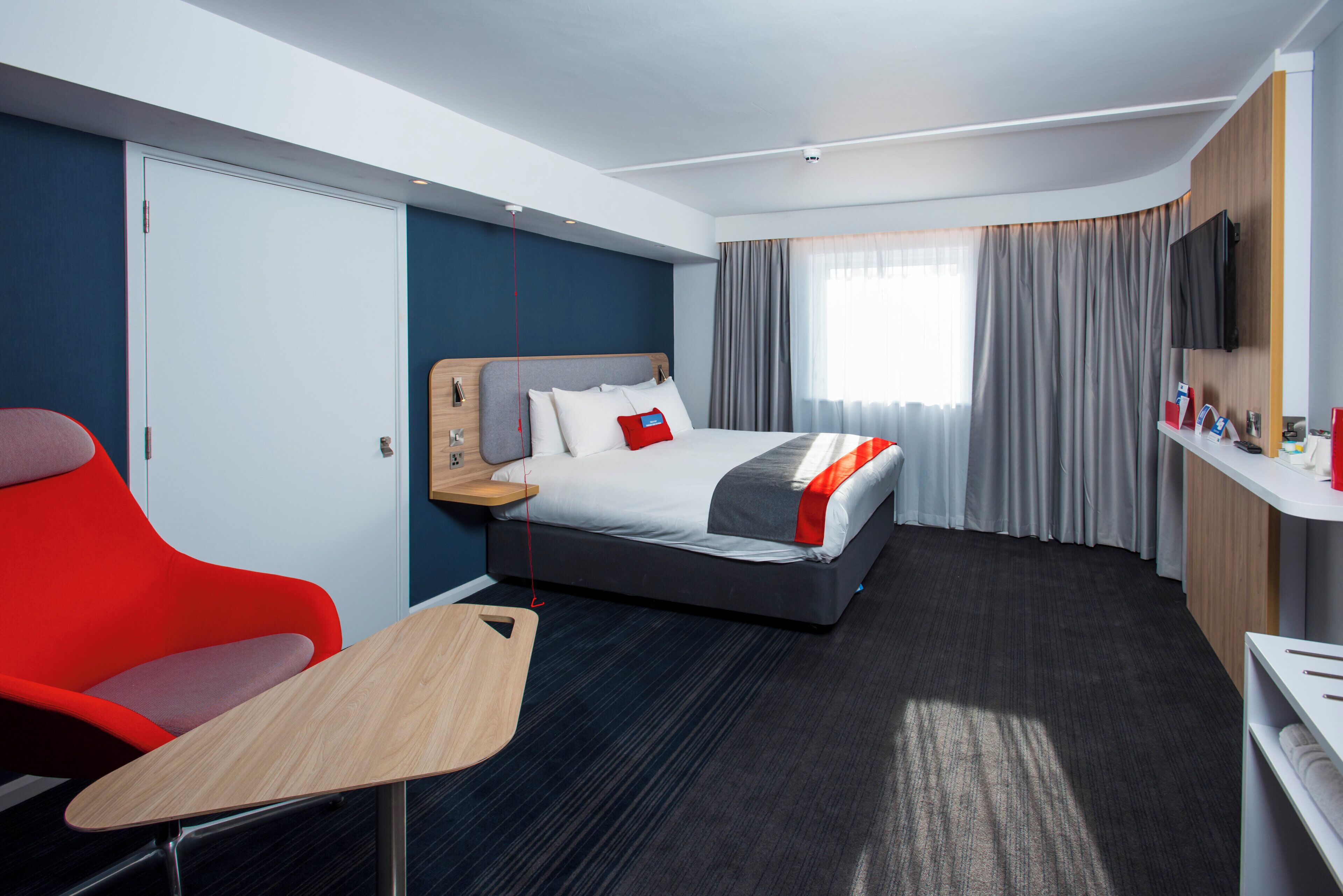 A hotel room with a bed, a desk, a chair, a TV, and a window with curtains.