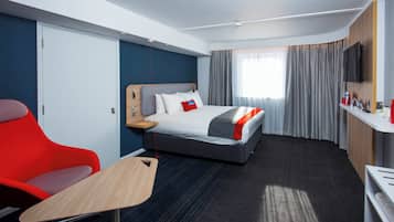 Una habitación de hotel con cama, escritorio, silla, televisión y ventana con cortinas.
