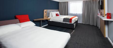 Habitación de hotel con una cama grande, una cama más pequeña, un escritorio, una silla, un televisor y una ventana con cortinas.