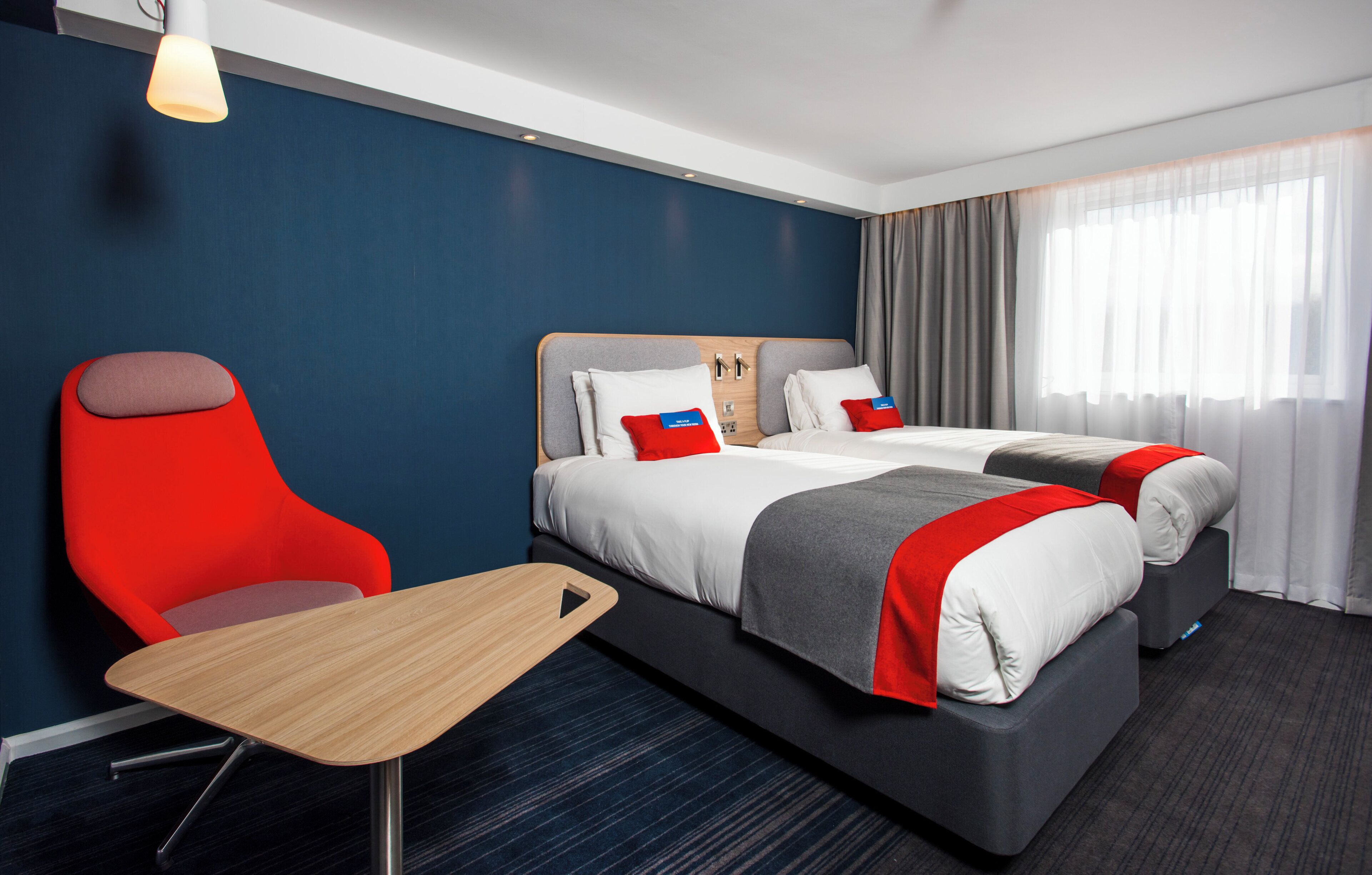 Une chambre d’hôtel avec un lit, deux oreillers, une chaise, une petite table et une fenêtre avec des rideaux.