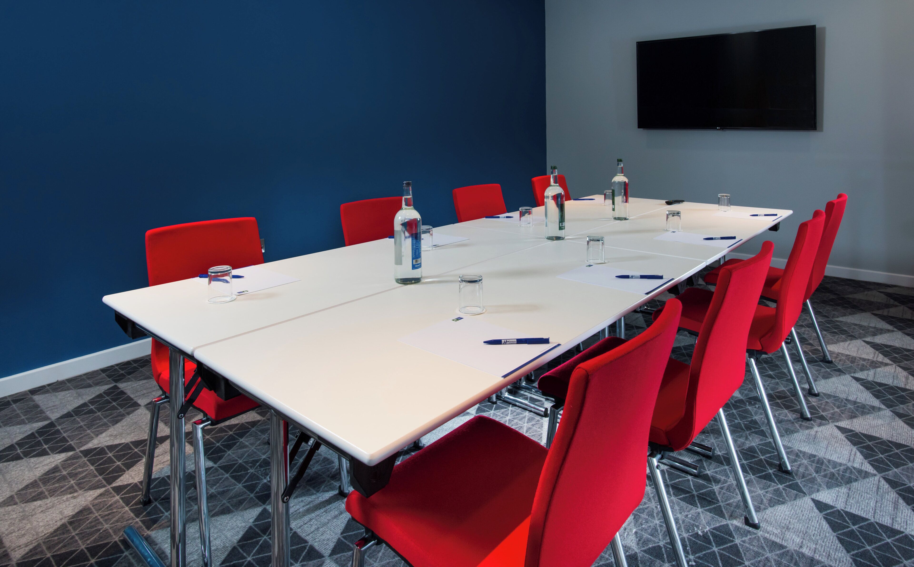 A conference room with a long table, red chairs, and a flat-screen TV on the wall.