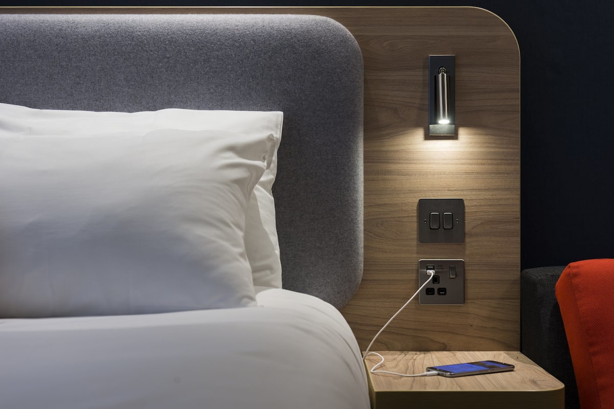 A neatly made bed with white linens, a bedside table with a smartphone charging, and a wall-mounted light fixture.