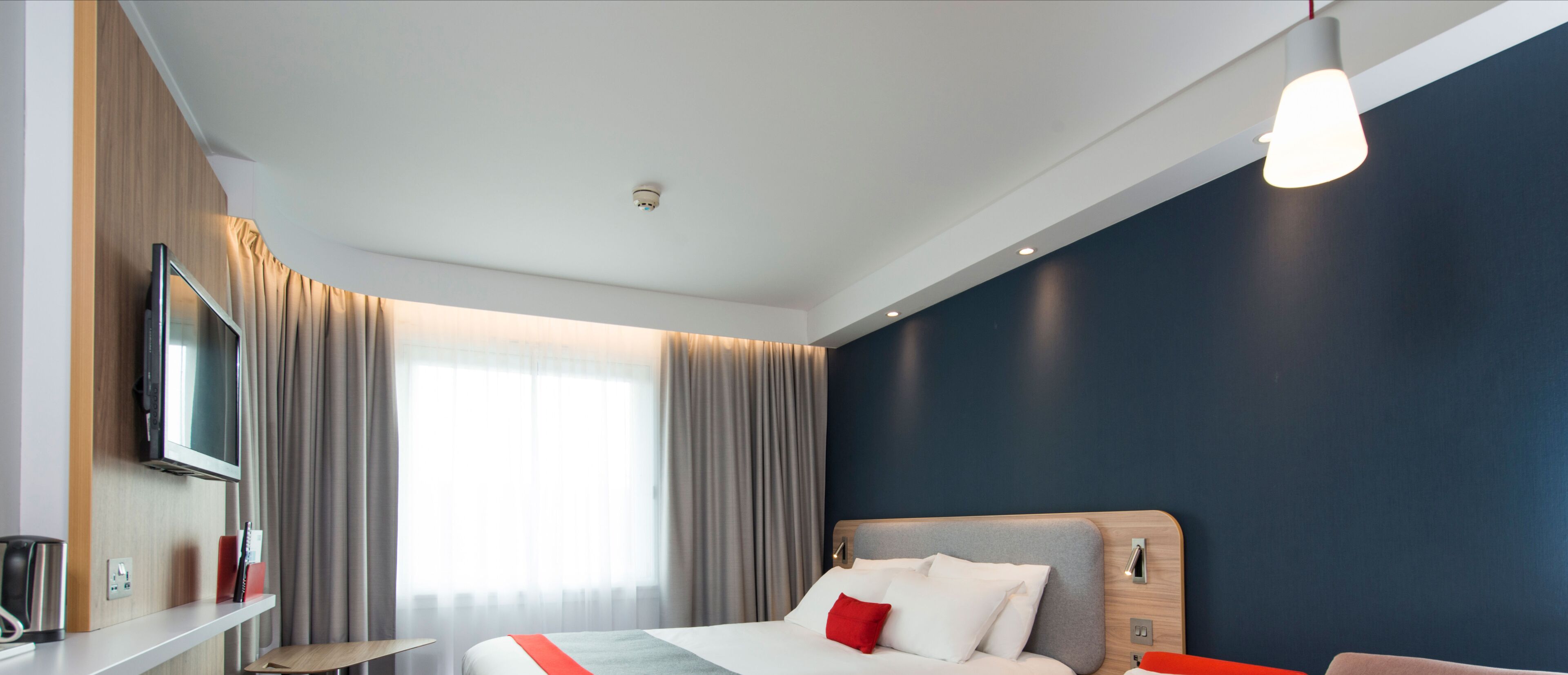 A hotel room with a bed, a TV mounted on the wall, a bedside table with a lamp, and a window with curtains.