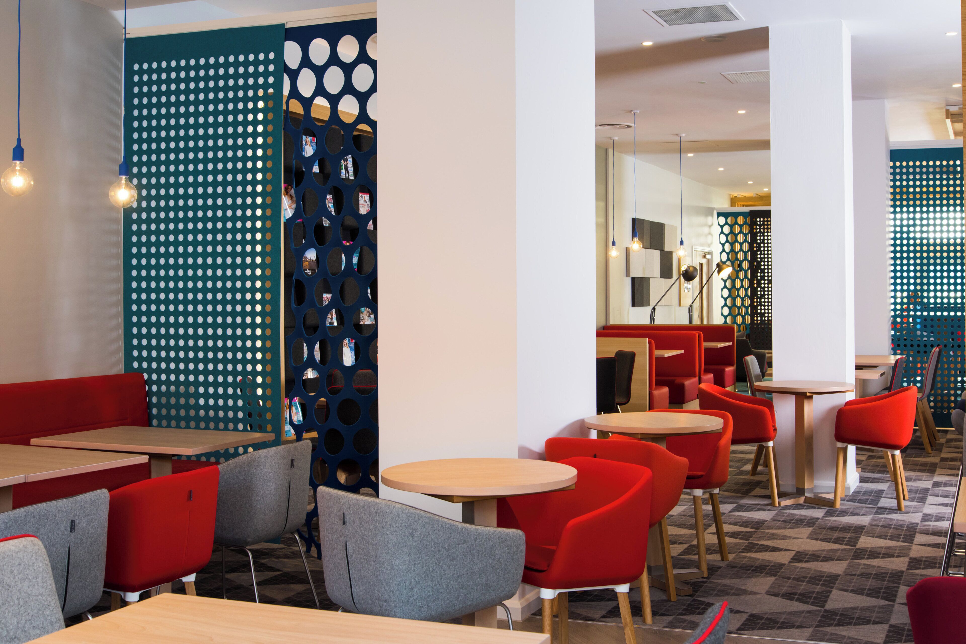 A modern interior with round tables and red and grey chairs, featuring a patterned wall and a blue partition with circular cutouts.