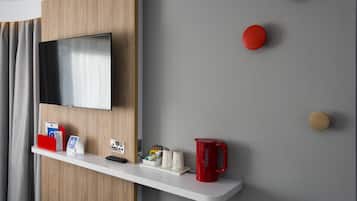 A modern hotel room with a mounted TV, a white shelf with various items, and a grey wall with wooden accents.