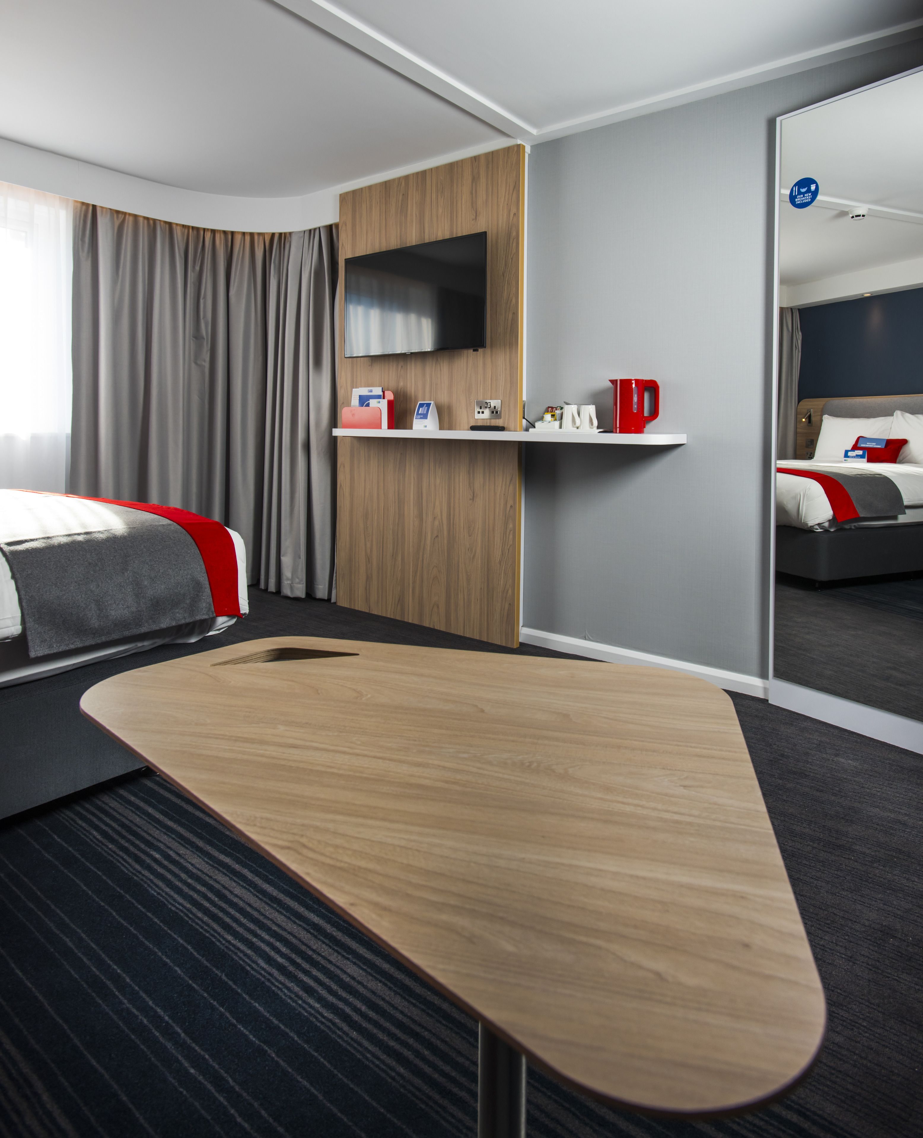 Una habitación de hotel moderna con un escritorio de madera, un televisor de pantalla plana, un minibar y una cama con detalles en rojo.