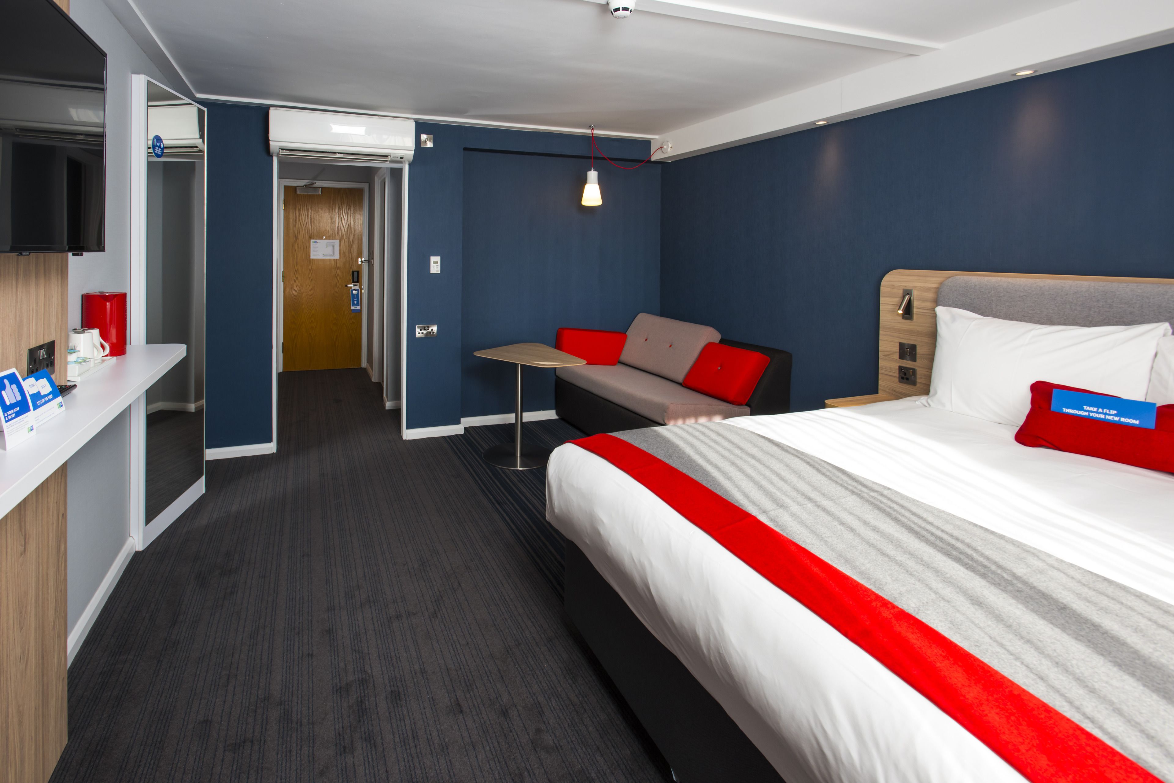 Quarto de hotel com uma cama, escrivaninha, cadeira e mesa pequena.