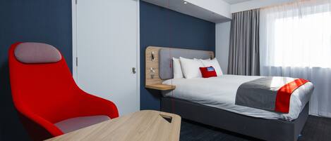 Une chambre d’hôtel avec un lit, une chaise rouge, une table en bois et une fenêtre avec des rideaux.