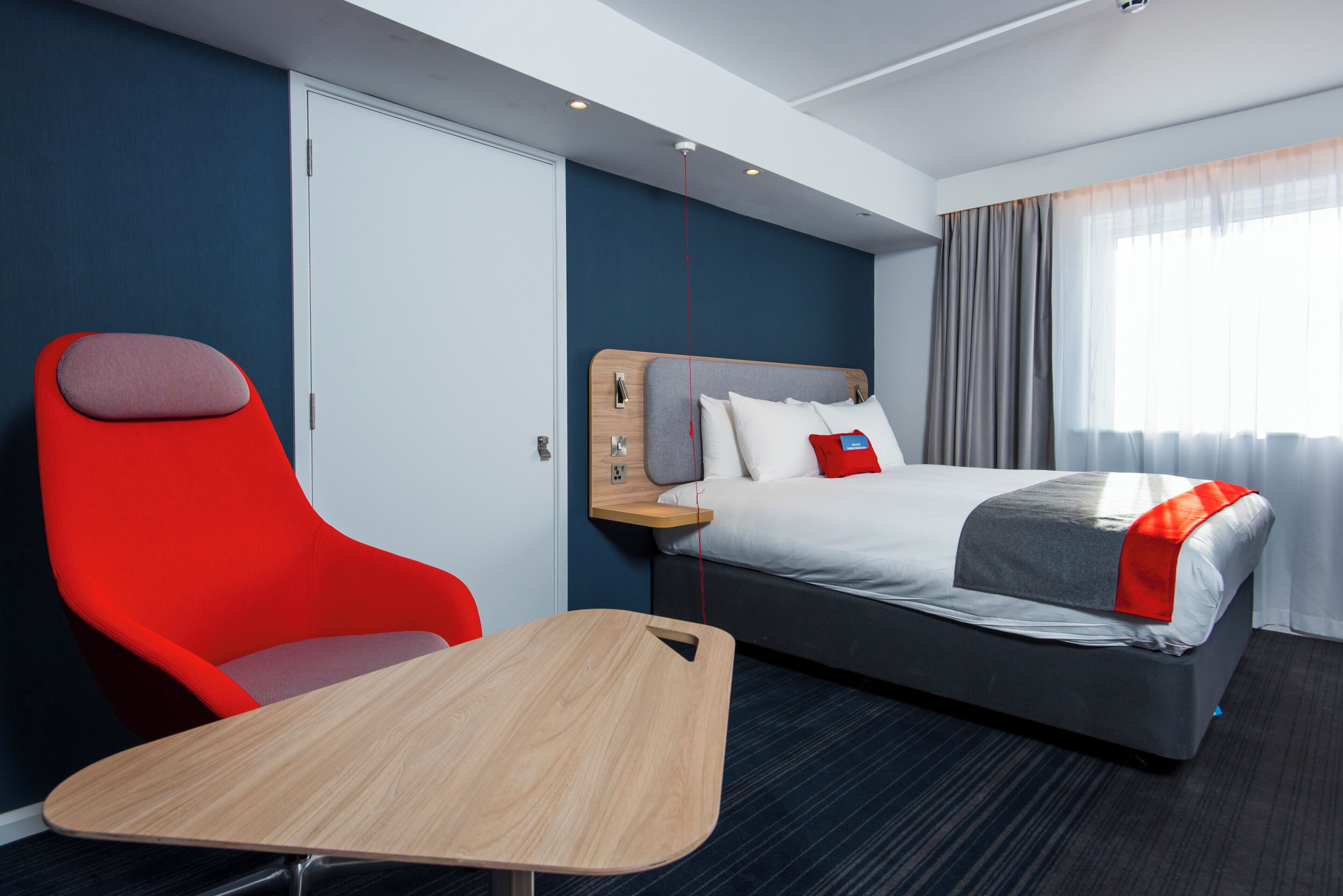 Una habitación de hotel con una cama, una silla roja, una mesa de madera y vistas al exterior.