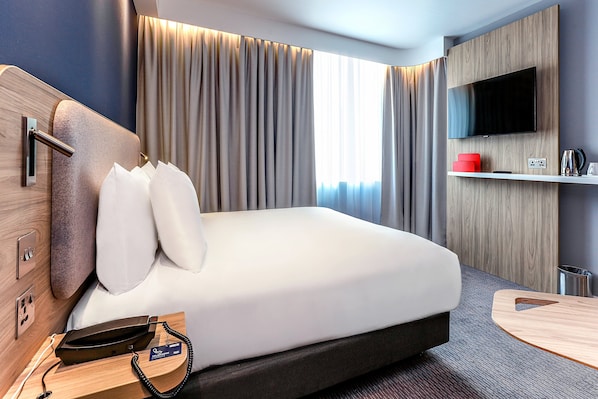 Una habitación de hotel moderna con una cama grande, un televisor de pantalla plana montado en un panel de madera, una mesita de noche con un teléfono y cargador, y una ventana con cortinas.