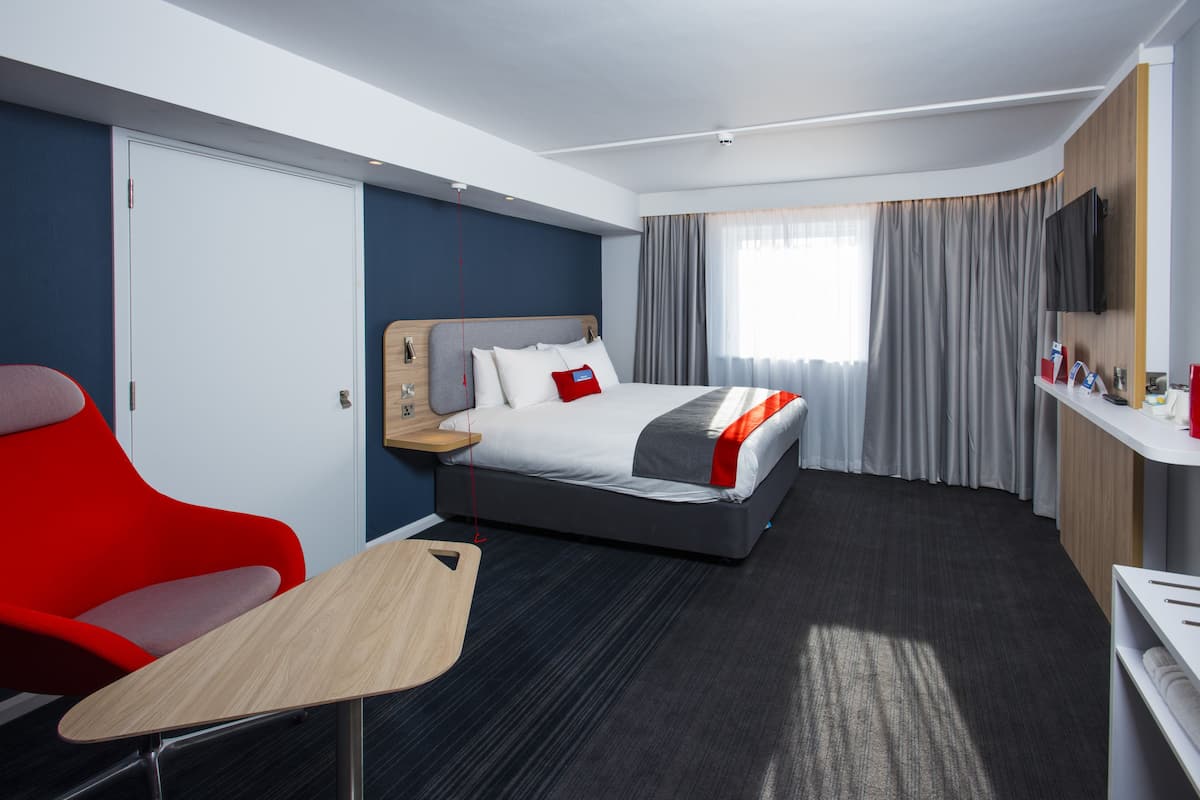 A hotel room with a bed, a red chair, a small table, a TV, and a window with curtains.