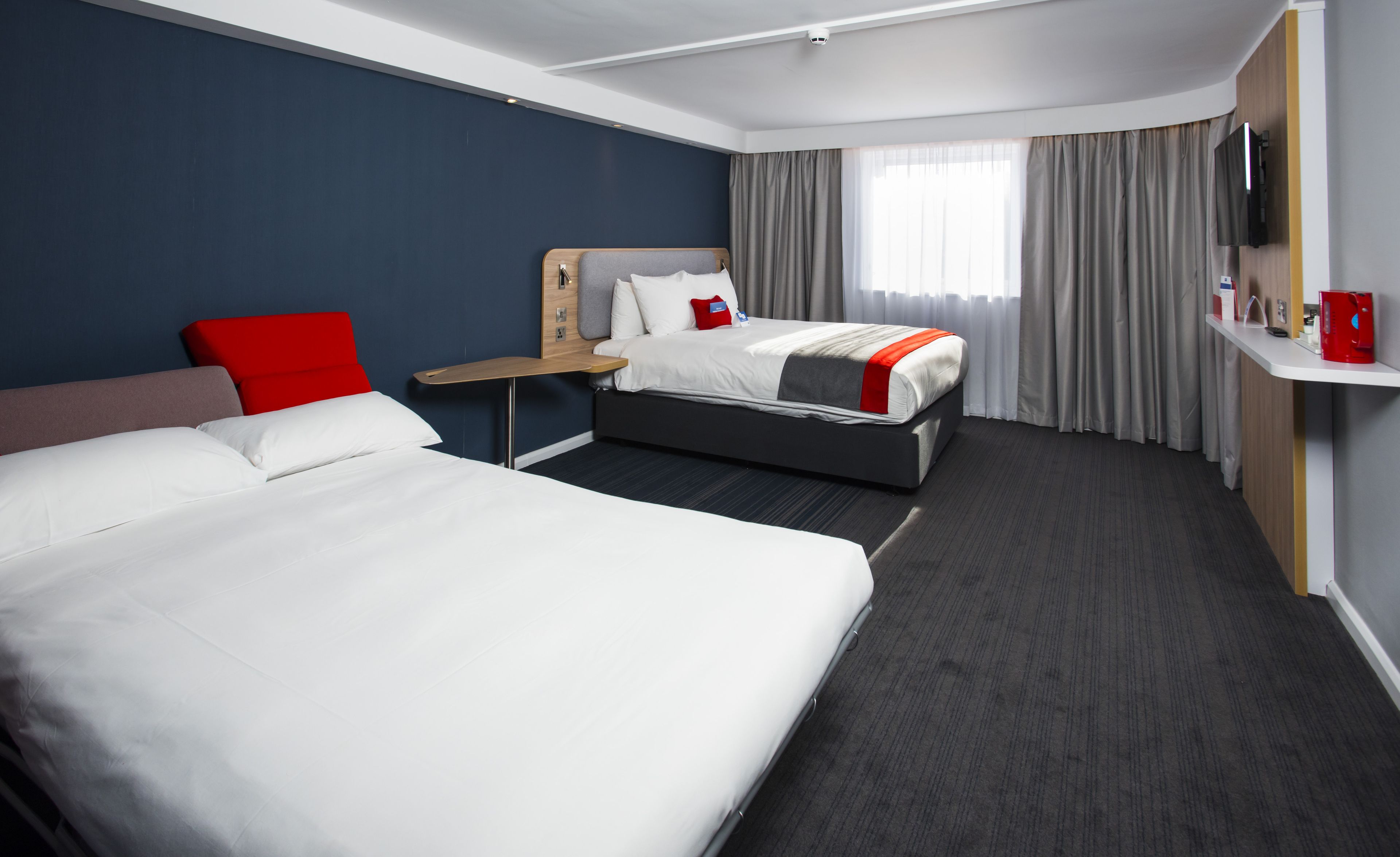 A hotel room with a large bed, a smaller bed, a desk, a chair, a TV, and a window with curtains.