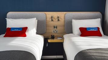 Habitación de hotel con dos camas, cada una con un almohadón y una etiqueta que dice "Take a flip through your new room". Entre las camas hay una mesita de noche de madera con un teléfono y un bolígrafo.