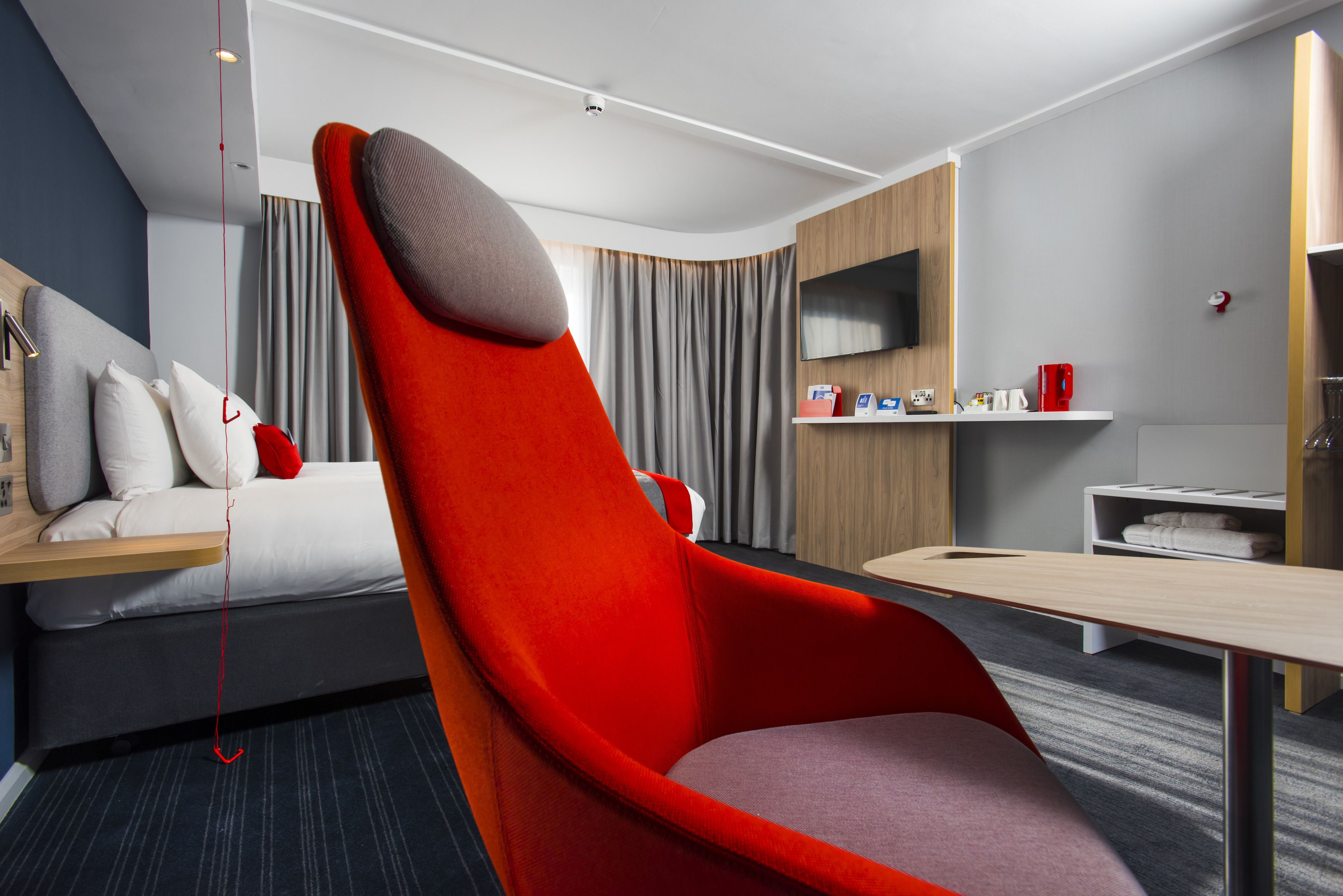 Una habitación de hotel moderna con una cama grande, un sillón rojo, un televisor de pantalla plana, un estante de madera con artículos de aseo y un escritorio con una silla.